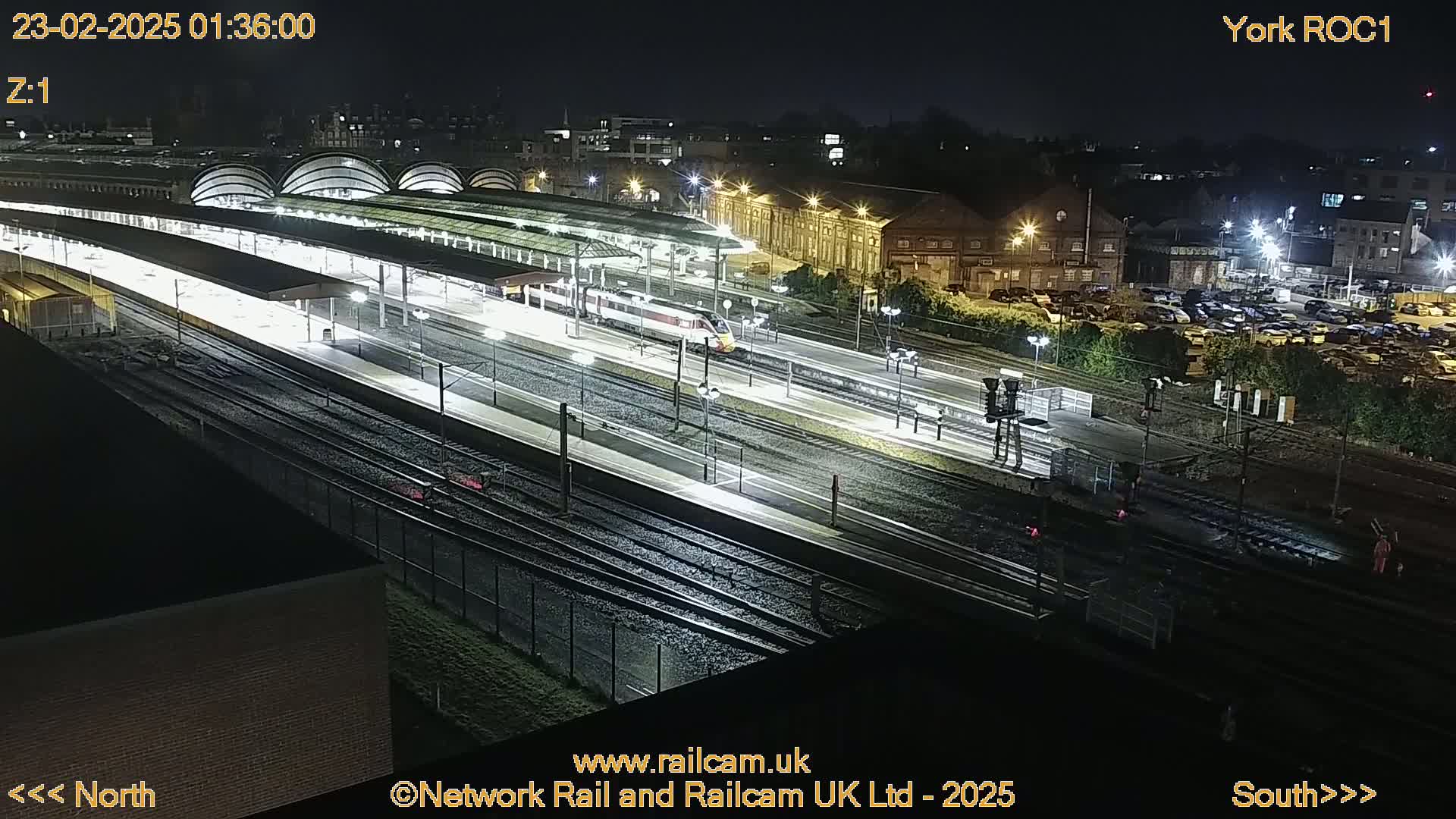A high-angle nighttime view of a train station with a train on the tracks, illuminated by artificial lights.