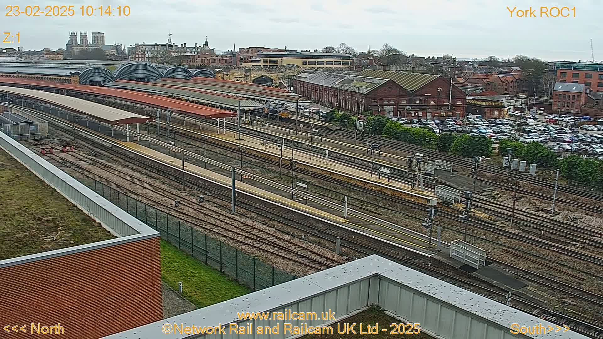 A high-angle view of a train station with numerous tracks and platforms, under an overcast sky, showing a large parking lot and a city in the background.