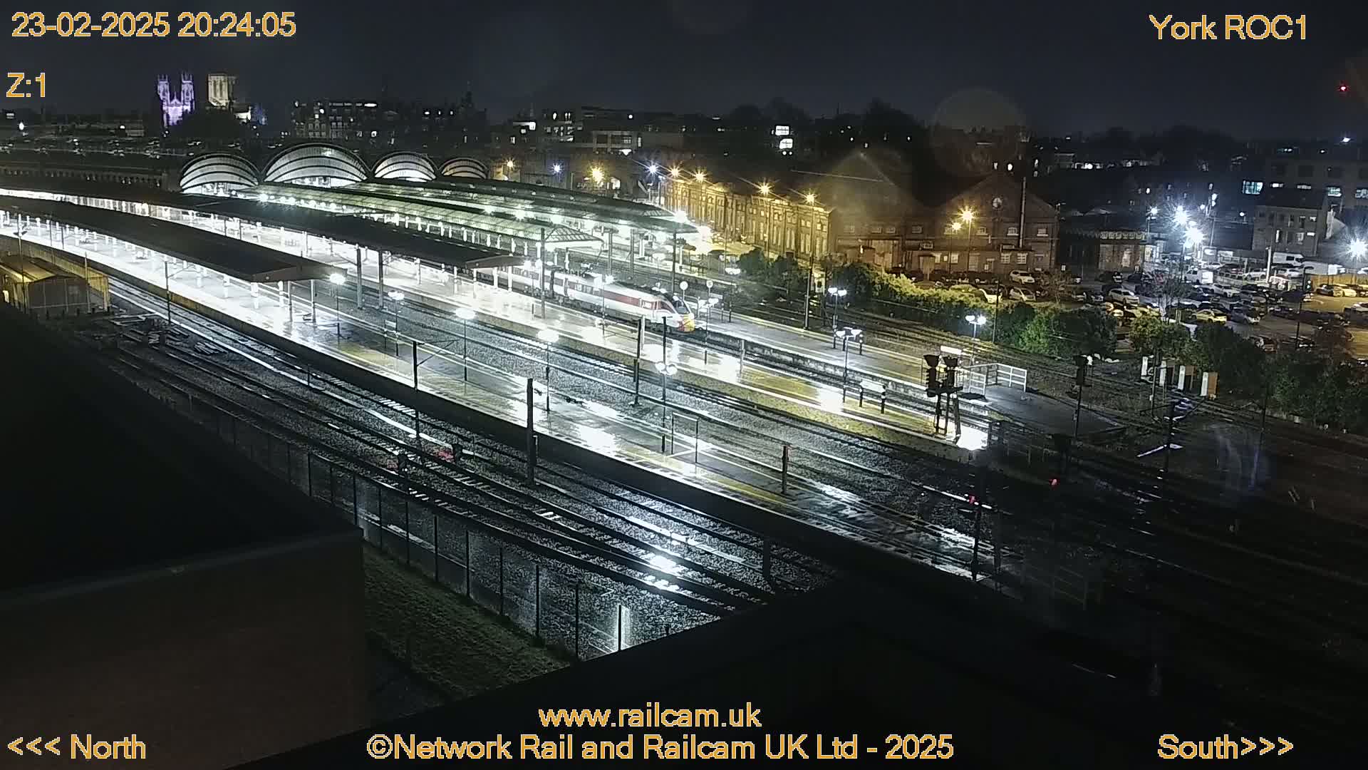 A rain-slicked train station at night, with a train on the tracks and numerous platforms under covered walkways.