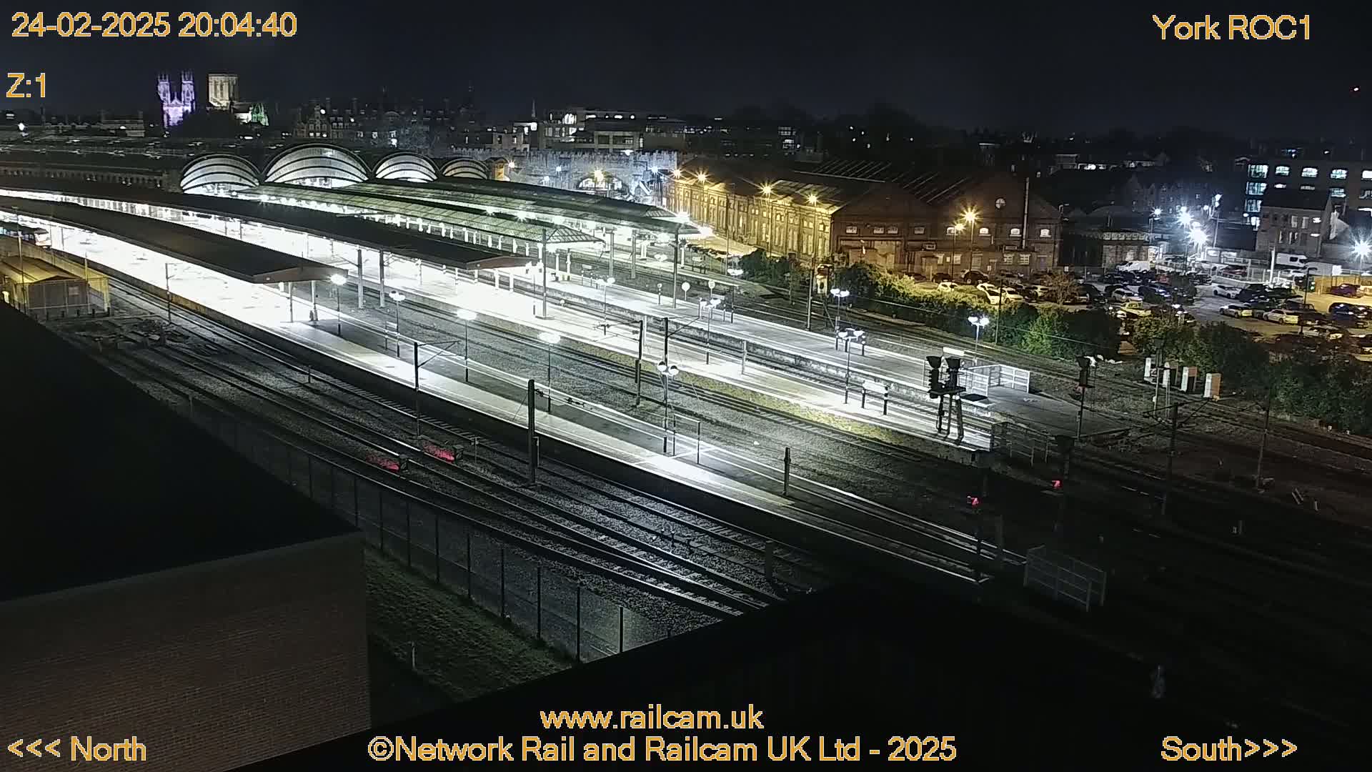 A nighttime aerial view of a brightly lit train station with numerous tracks and platforms, surrounded by buildings and a parking lot.