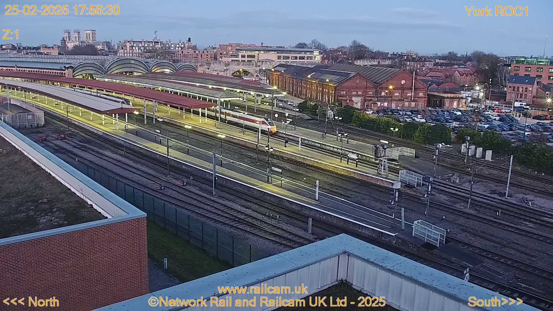A high-angle, twilight view of a train station with a high-speed train on the tracks, surrounded by railway lines, buildings, and a parking lot.