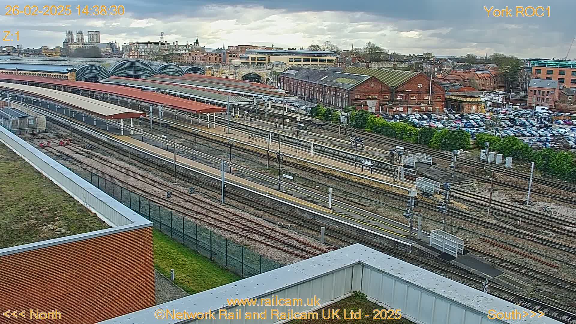 A wide view of a railway station and surrounding buildings under a cloudy sky shows many train tracks, platforms, and a large parking lot.