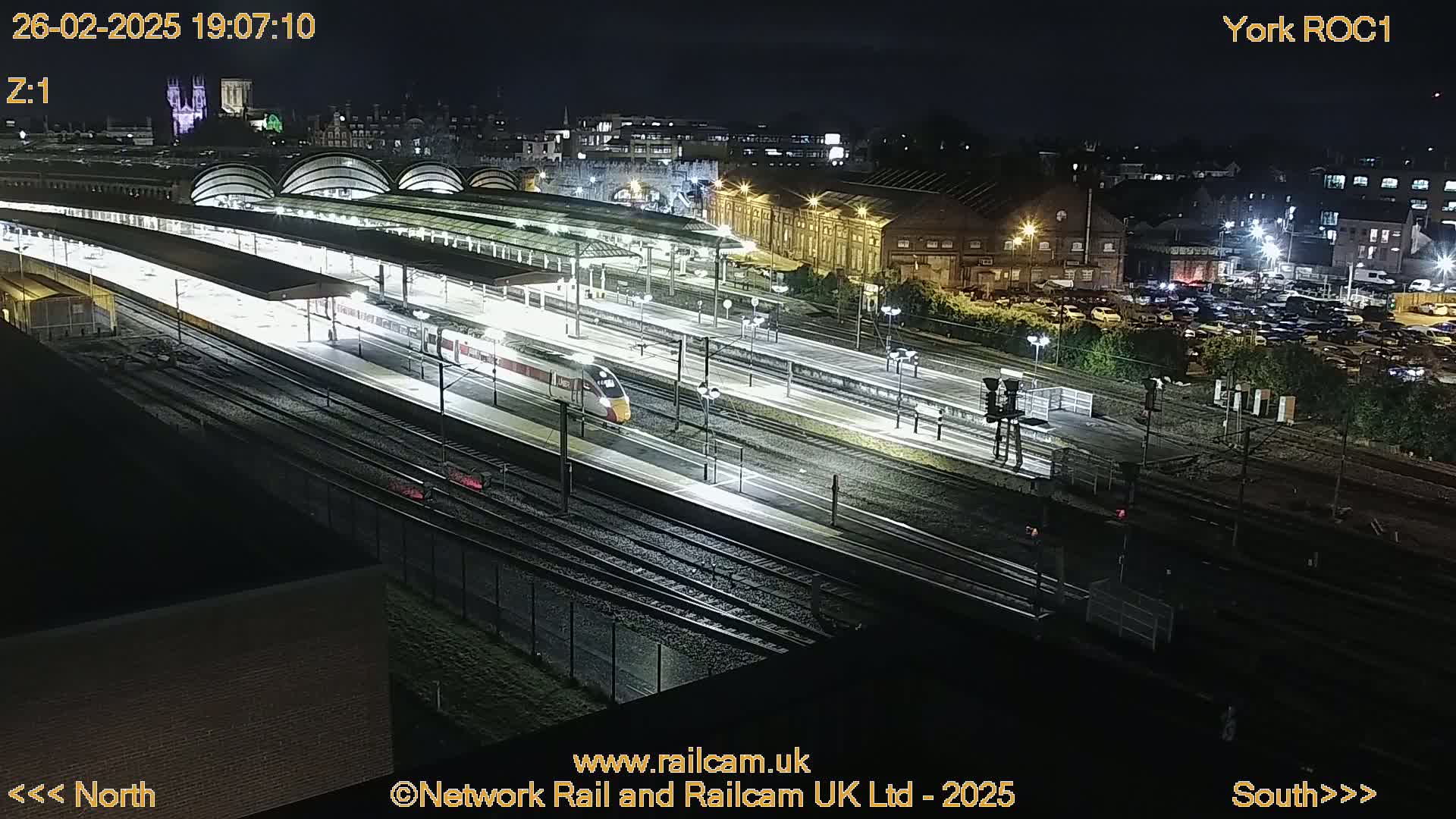 A high-angle nighttime view of a train station with a high-speed train passing through, illuminated by artificial lights.