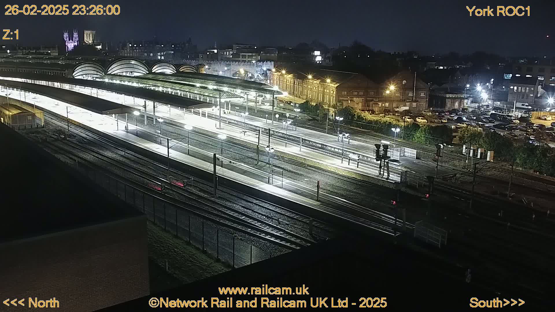 A nighttime, high-angle view of a brightly lit train station with numerous tracks and platforms, surrounded by dark buildings and a parking lot.