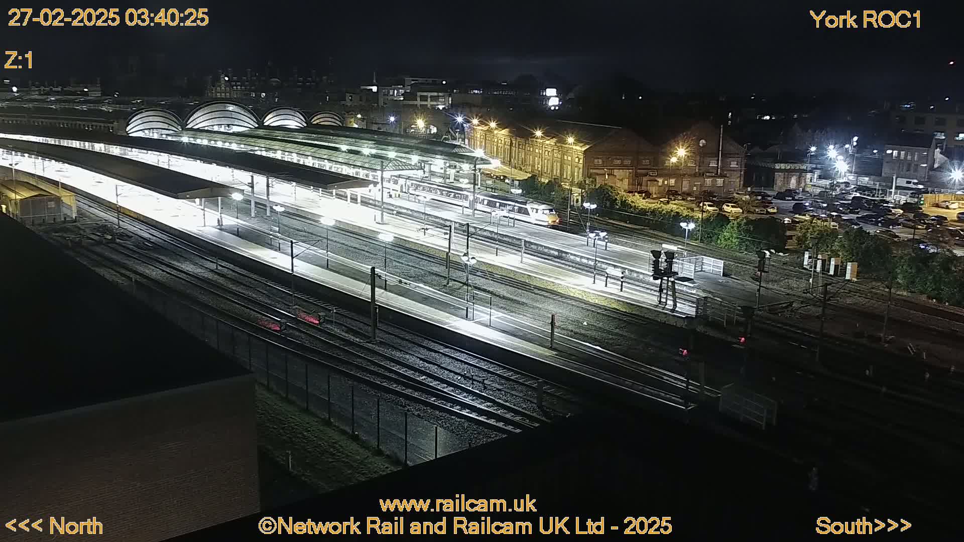 A nighttime aerial view of a train station with a train departing and a parking lot visible beyond the tracks.
