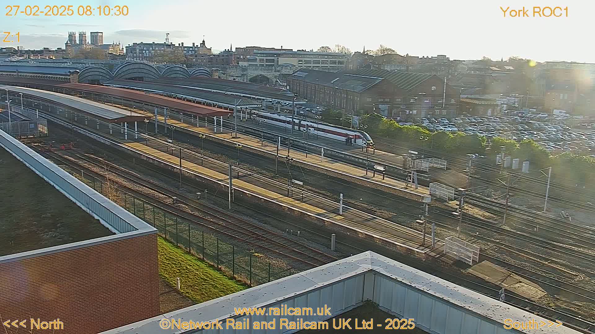 A high-angle, long shot of a train station on a sunny day, showing a train on the tracks and numerous railway lines.