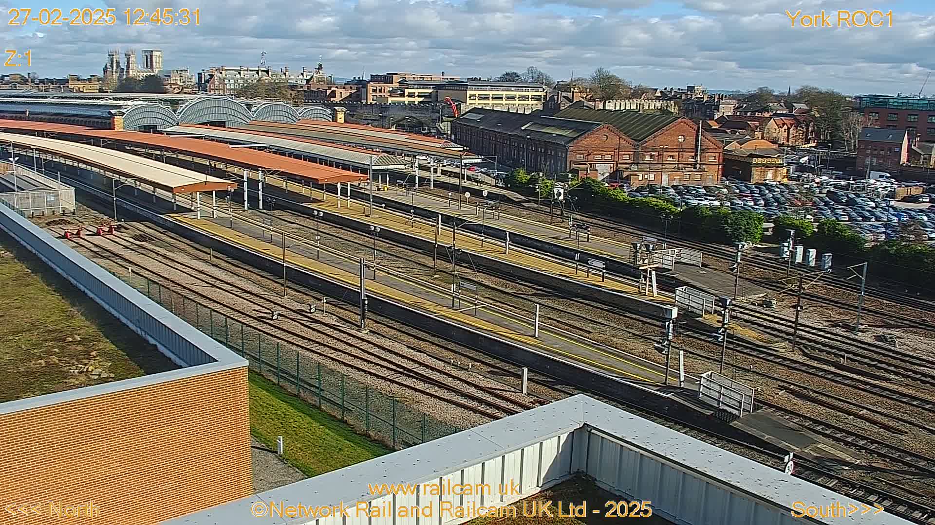 A high-angle, sunny view of a train station with many tracks, platforms, and a large parking lot, surrounded by buildings.