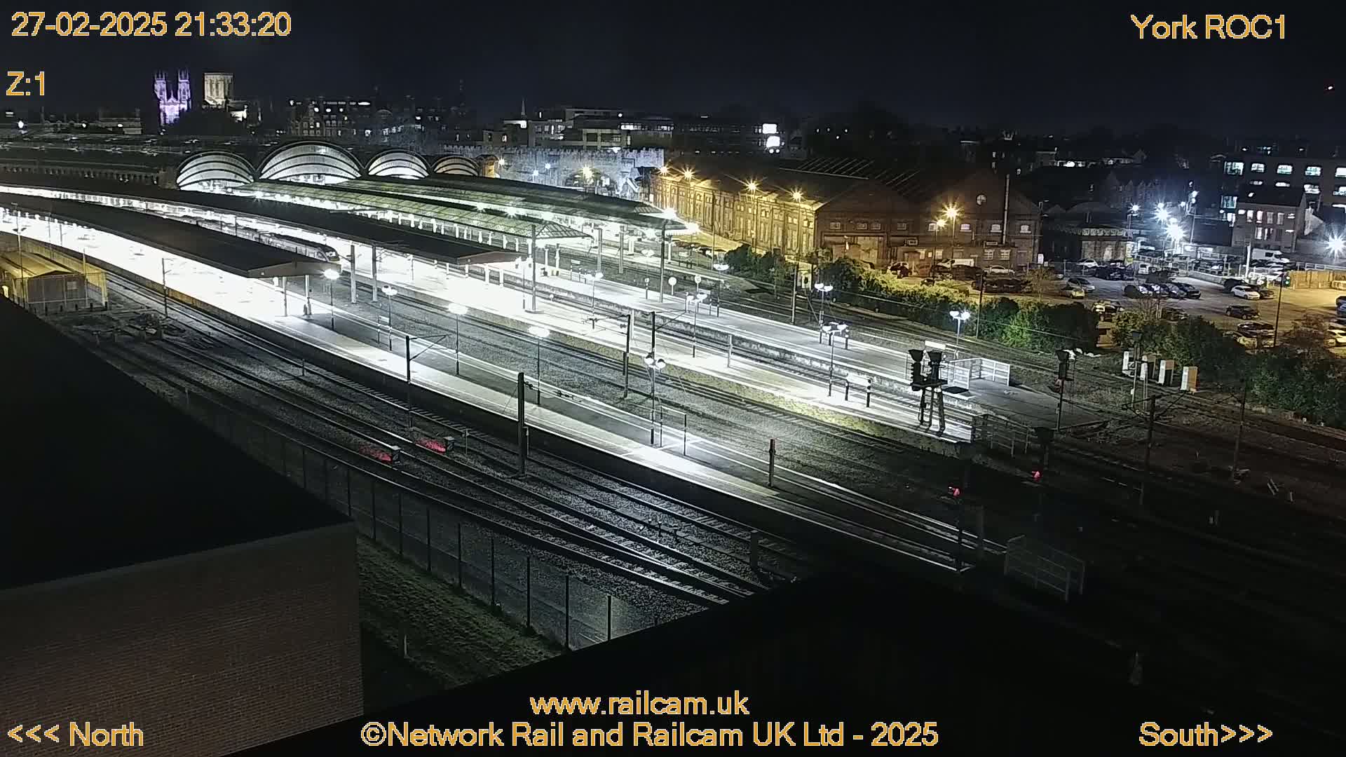 A nighttime, high-angle view of a train station with multiple tracks and platforms illuminated by artificial lights.