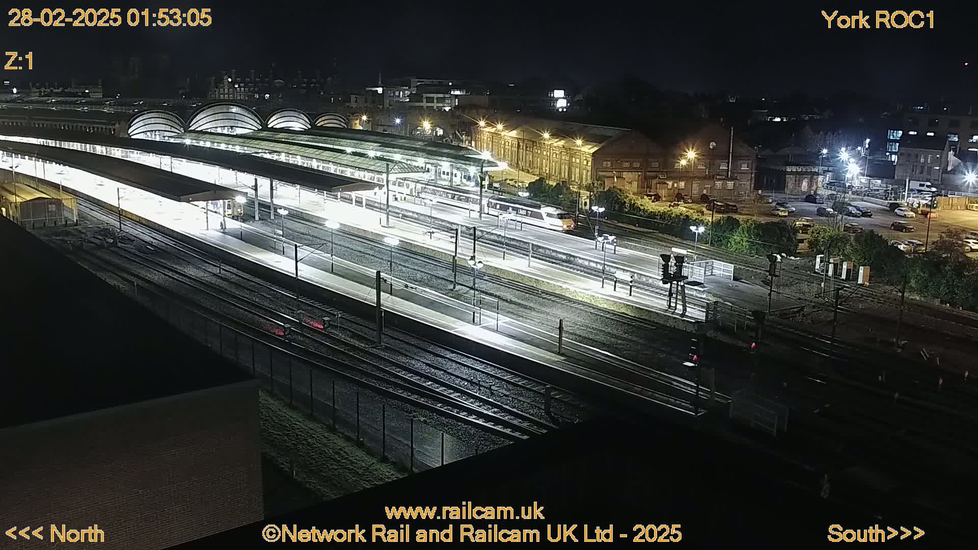 A brightly lit train station at night with a train departing from a platform, surrounded by tracks and buildings.