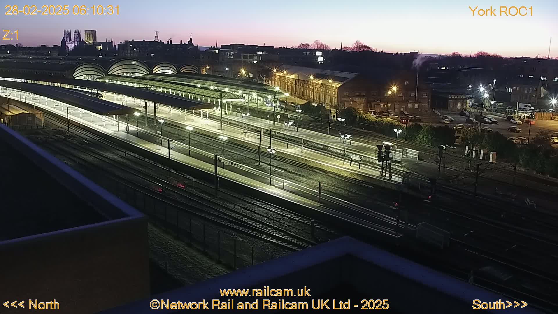 A nighttime, twilight view of a brightly lit train station with many tracks and platforms, situated near a city skyline.