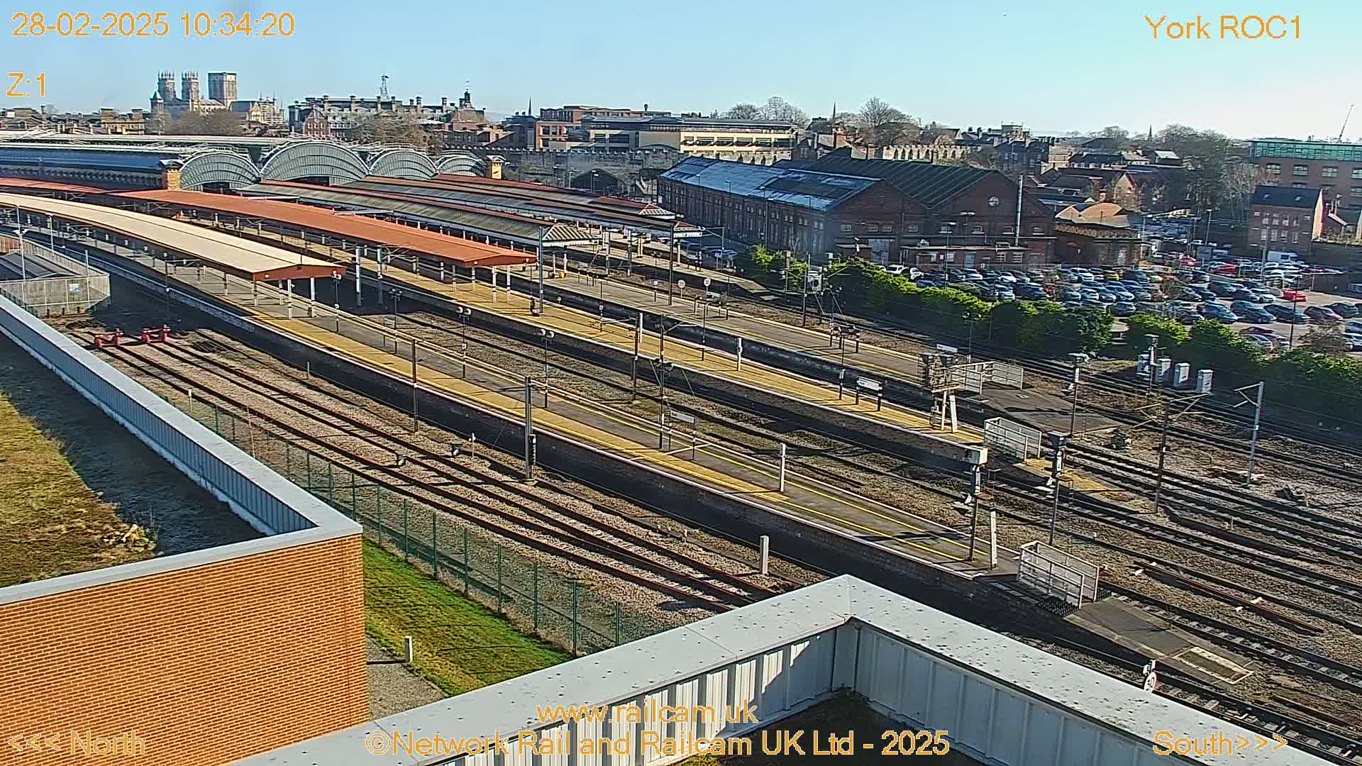 A high-angle view of a train station on a sunny day, showing numerous train tracks, platforms, and a large parking lot filled with cars.