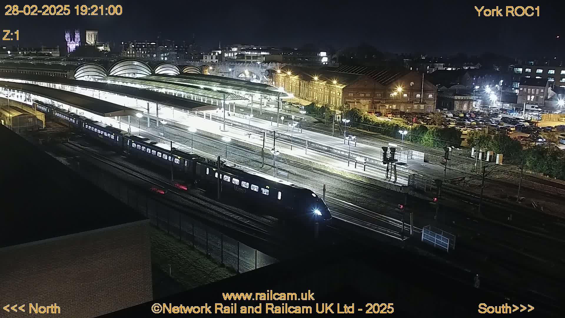 A high-angle nighttime view shows a train moving through a well-lit train station with multiple tracks, platforms, and surrounding buildings.