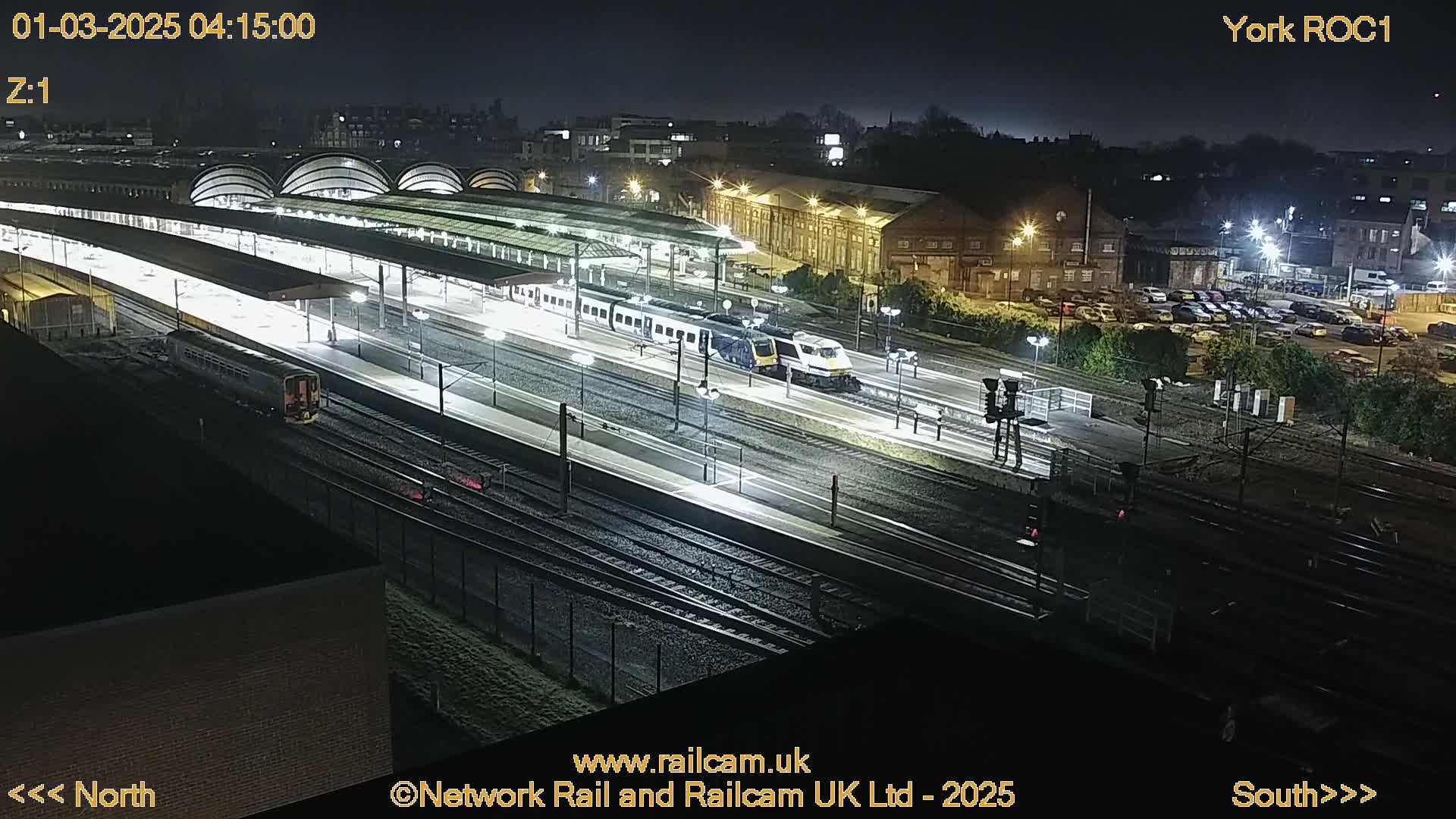 At night, several trains are at a brightly lit train station with multiple tracks.