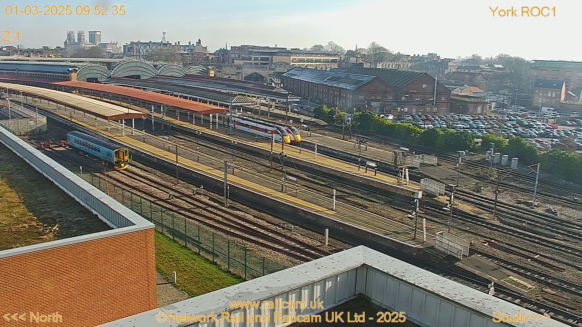 A high-angle, sunny view of a railway station with multiple train tracks, platforms, and several trains, including a light blue train and two high-speed trains.
