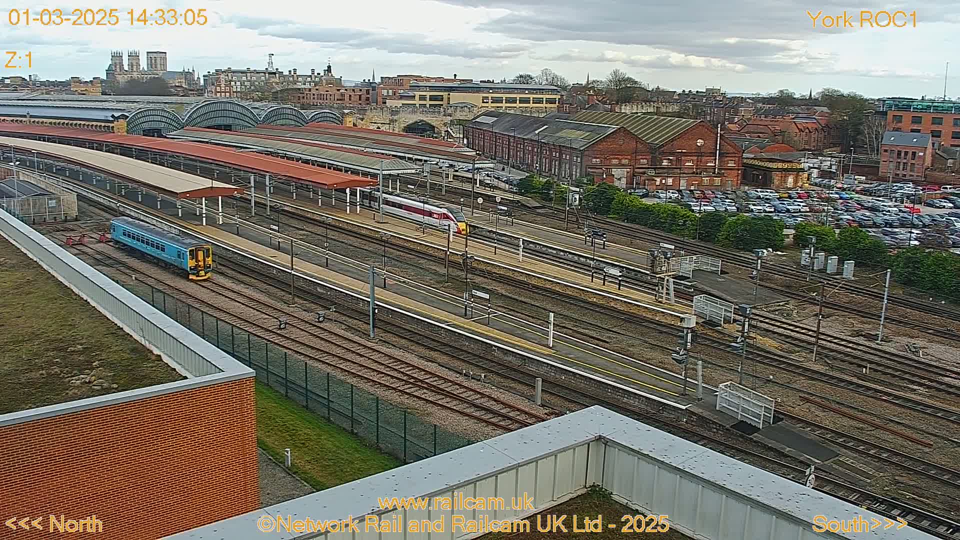 A high-angle view of a train station on an overcast day, showing multiple train tracks, platforms with trains, and a large parking lot, with a city and cathedral in the background.