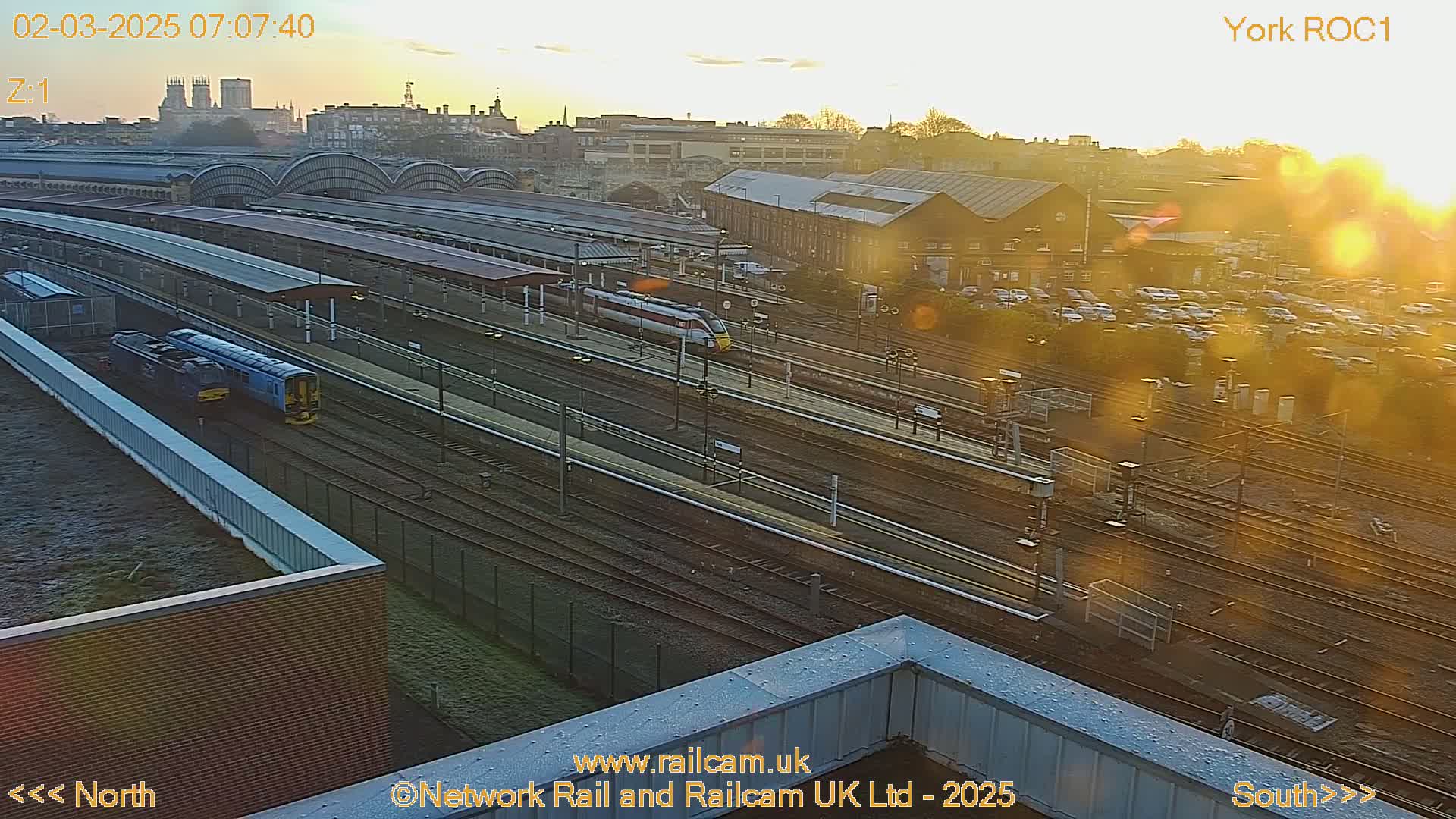 A high-angle, long shot of a train station at sunrise, showing multiple train tracks, several trains, and a city skyline in the background.