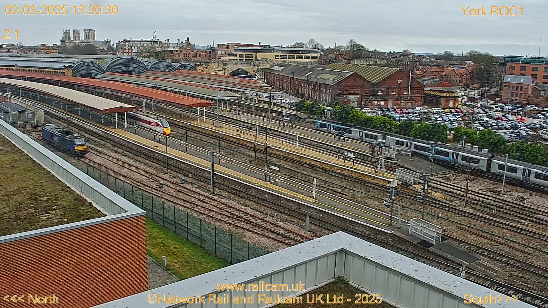 A high-angle view of a train station on an overcast day shows multiple train tracks with several trains, and a large parking lot in the background.