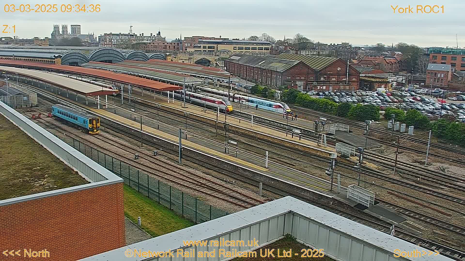 A high-angle view of a train station on an overcast day, showing multiple trains on various tracks, a large parking lot, and city buildings in the distance.