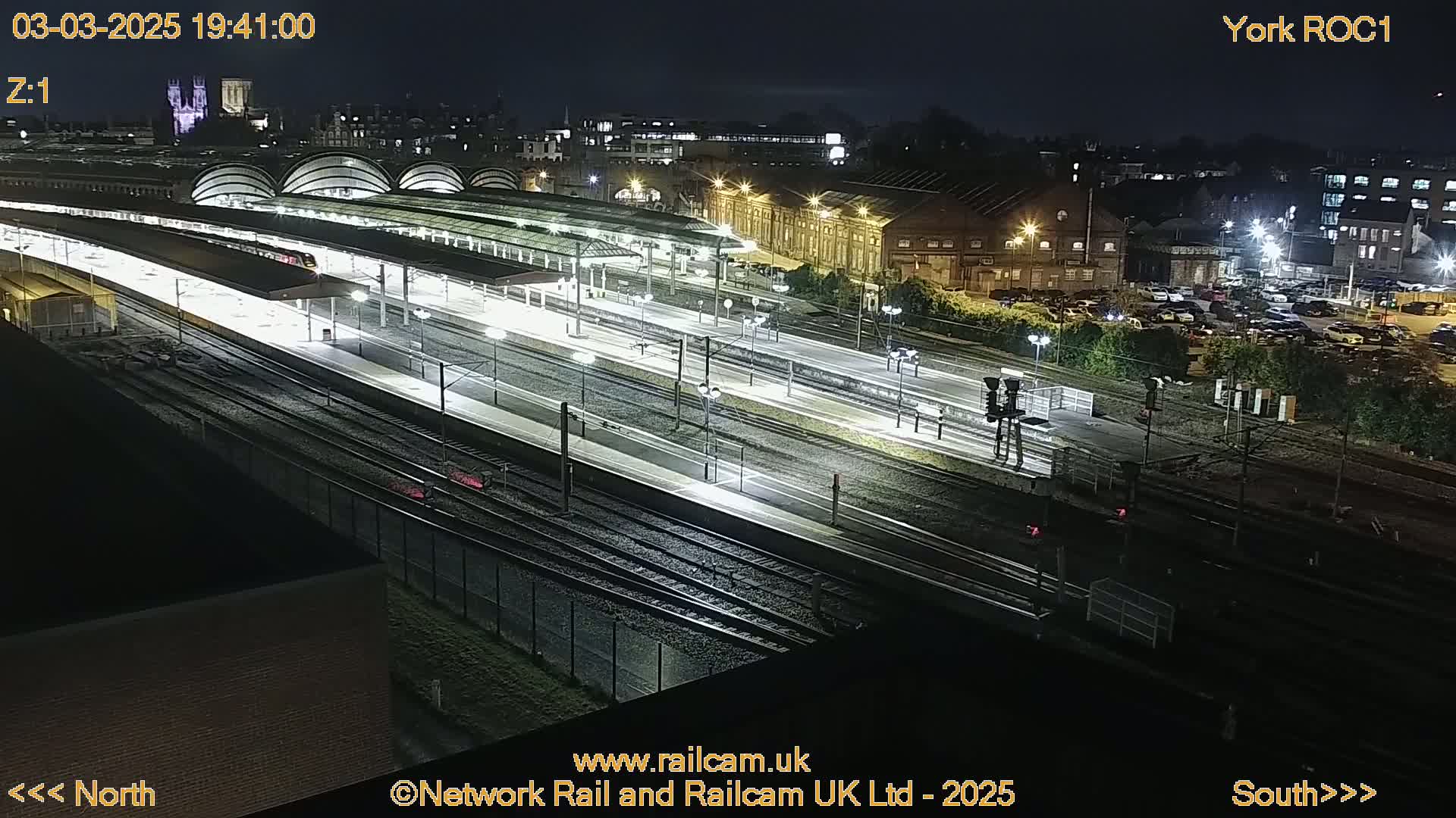 A nighttime aerial view of a brightly lit train station with multiple tracks and platforms, surrounded by buildings and a parking lot.