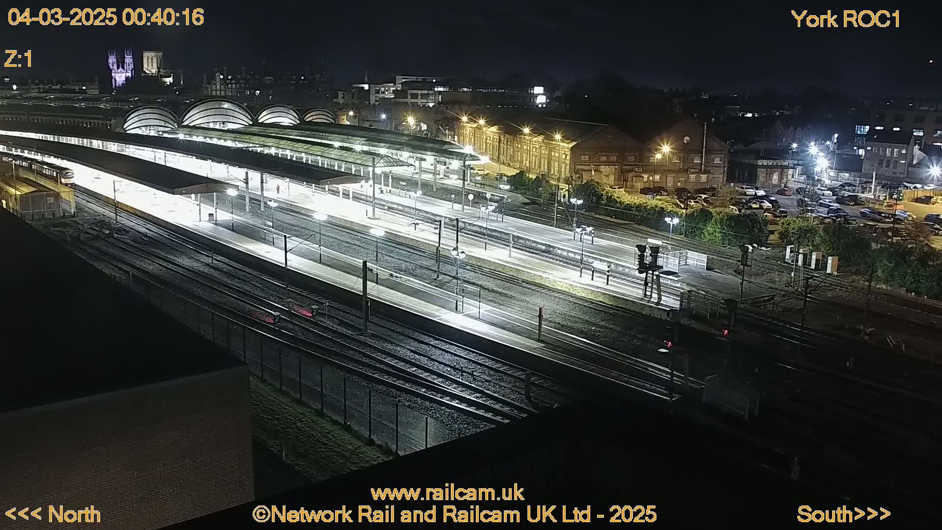 A nighttime, overhead view of a brightly lit train station with multiple tracks and platforms, surrounded by buildings and parking lots.