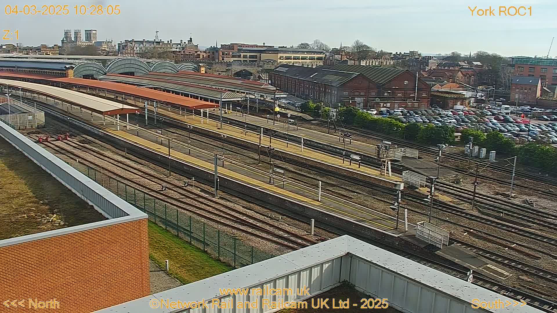 A high-angle view of a train station on a sunny day, showing numerous train tracks, platforms with covered waiting areas, and a large parking lot filled with cars.