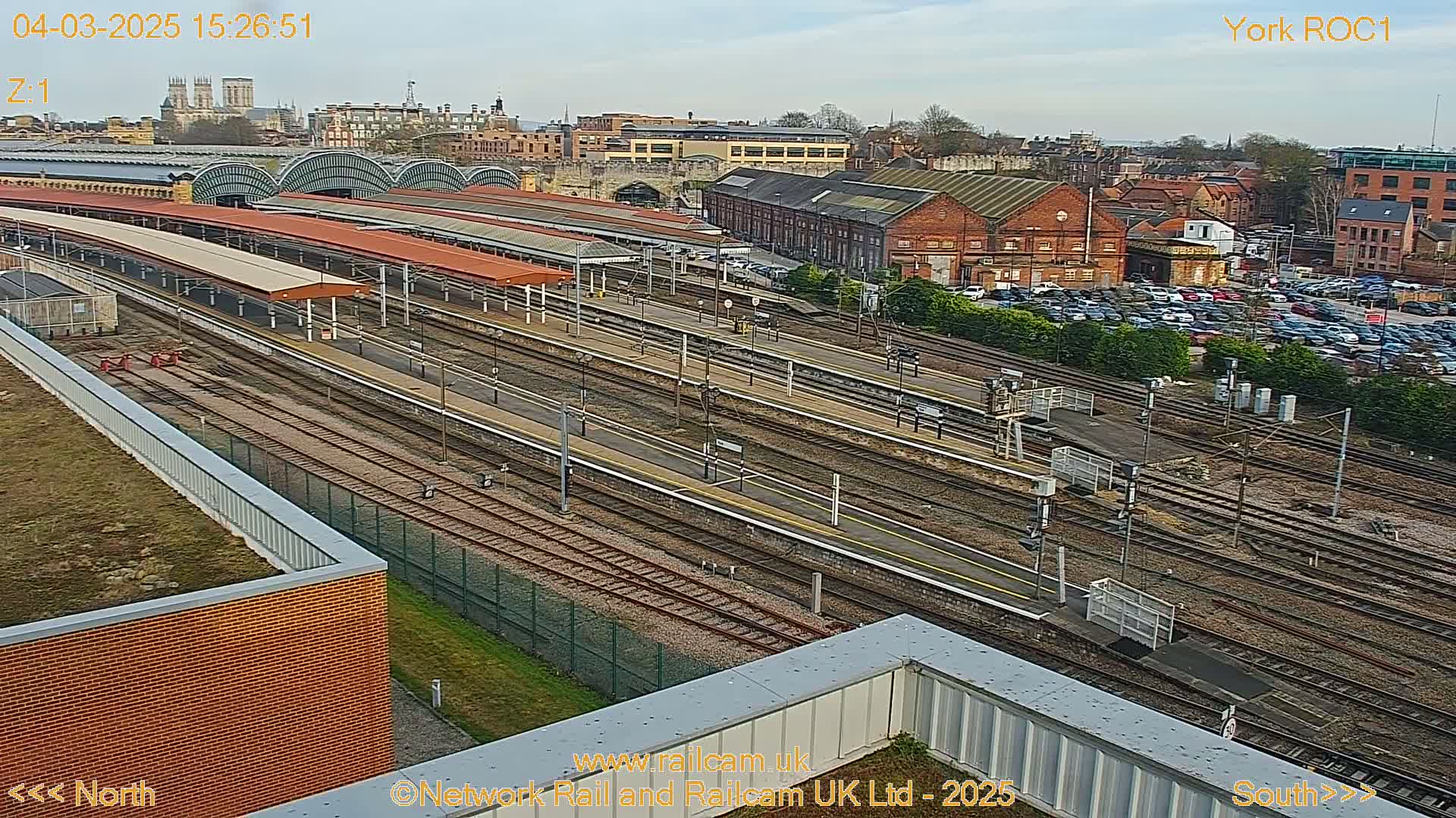 A high-angle, long shot of a railway station and surrounding area on a partly sunny day shows multiple train tracks, platforms with covered waiting areas, and a large parking lot filled with cars.