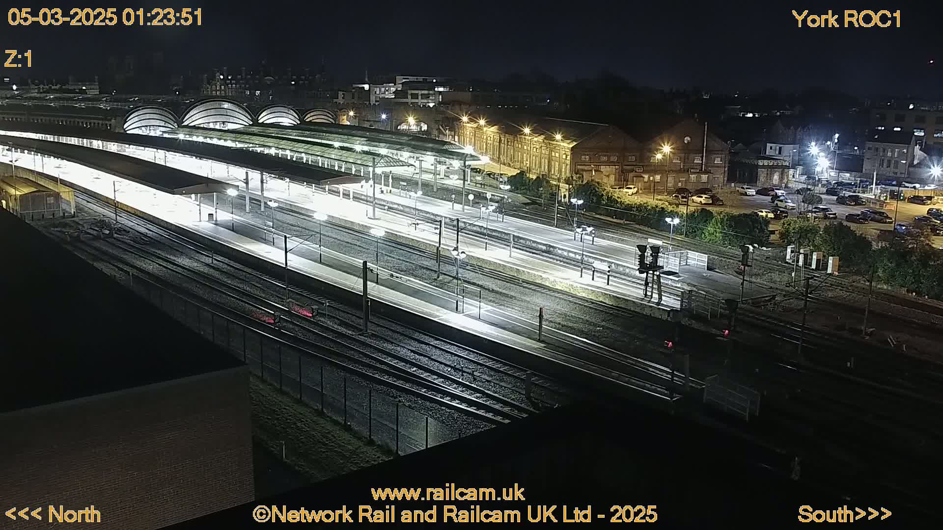 A nighttime, illuminated view of a train station with multiple tracks and platforms, surrounded by buildings and a parking lot.