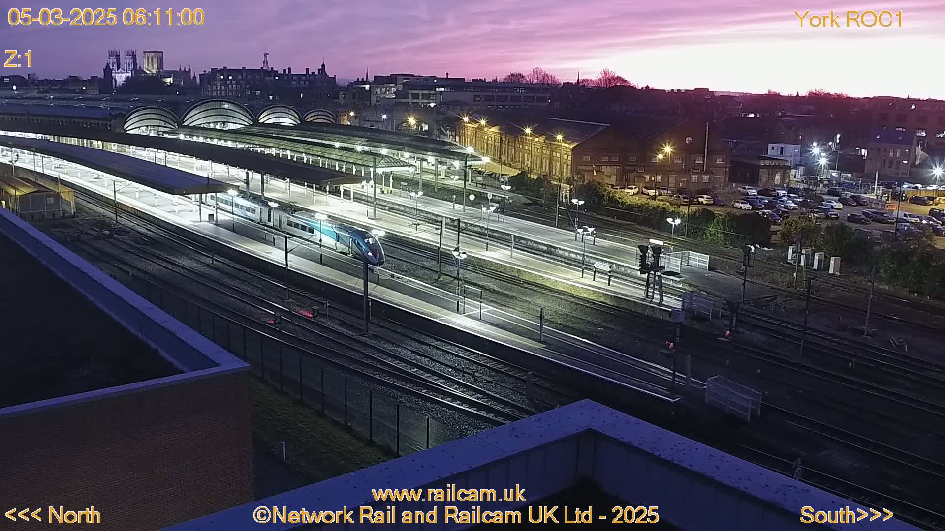 A high-angle, nighttime view of a train station with a train at a platform, under a purplish-pink twilight sky.