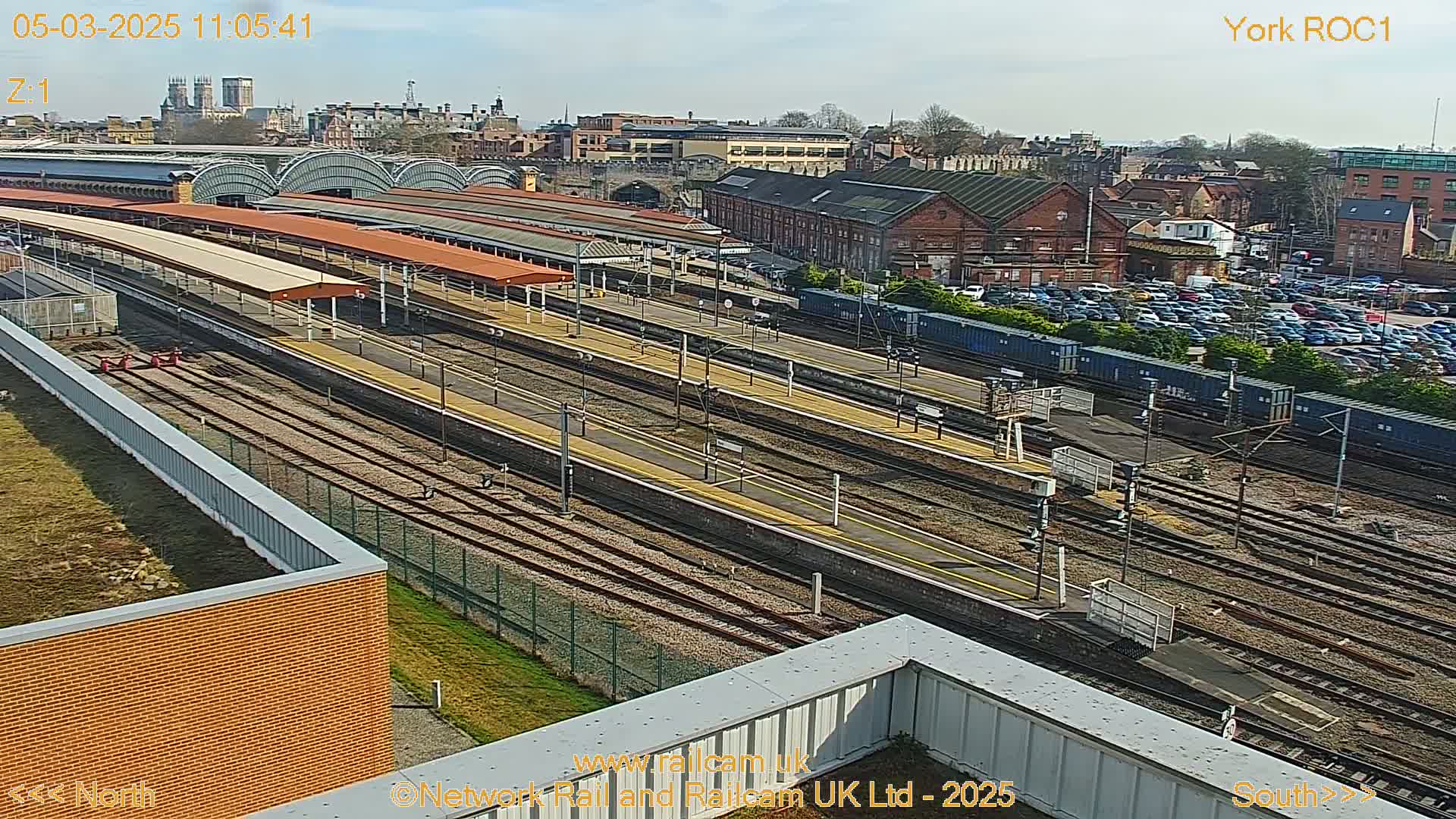 A high-angle, sunny view of a railway station with multiple tracks, platforms, and a large parking lot, with a city skyline in the background.