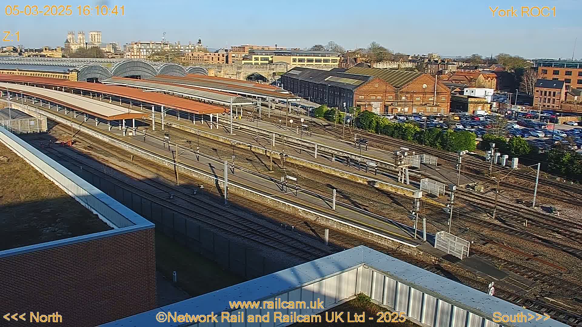 A high-angle, sunny view of a train station with numerous tracks, platforms, and surrounding buildings.