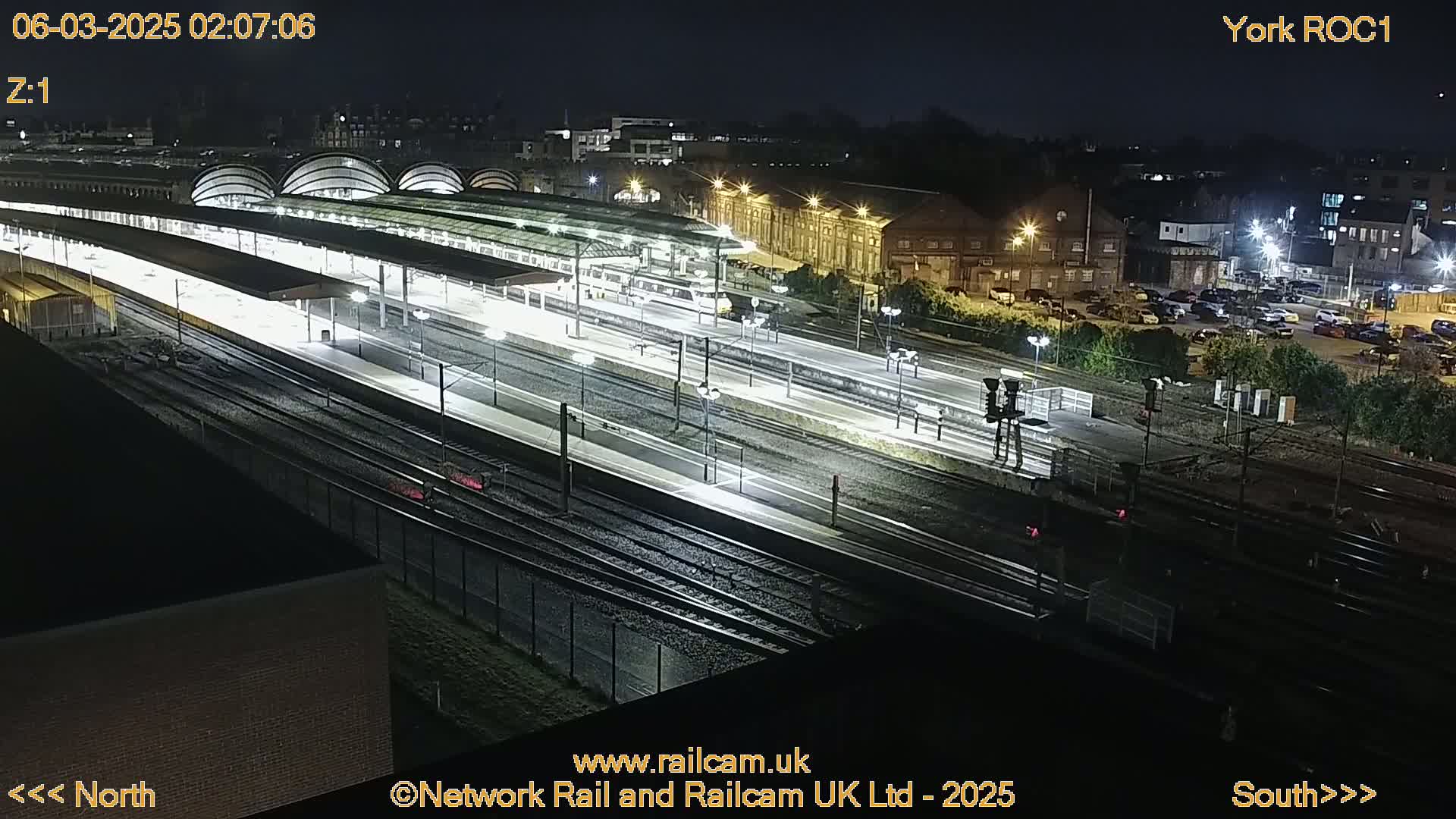 A nighttime, high-angle view of a brightly lit train station with multiple tracks and platforms, surrounded by buildings and parking lots.
