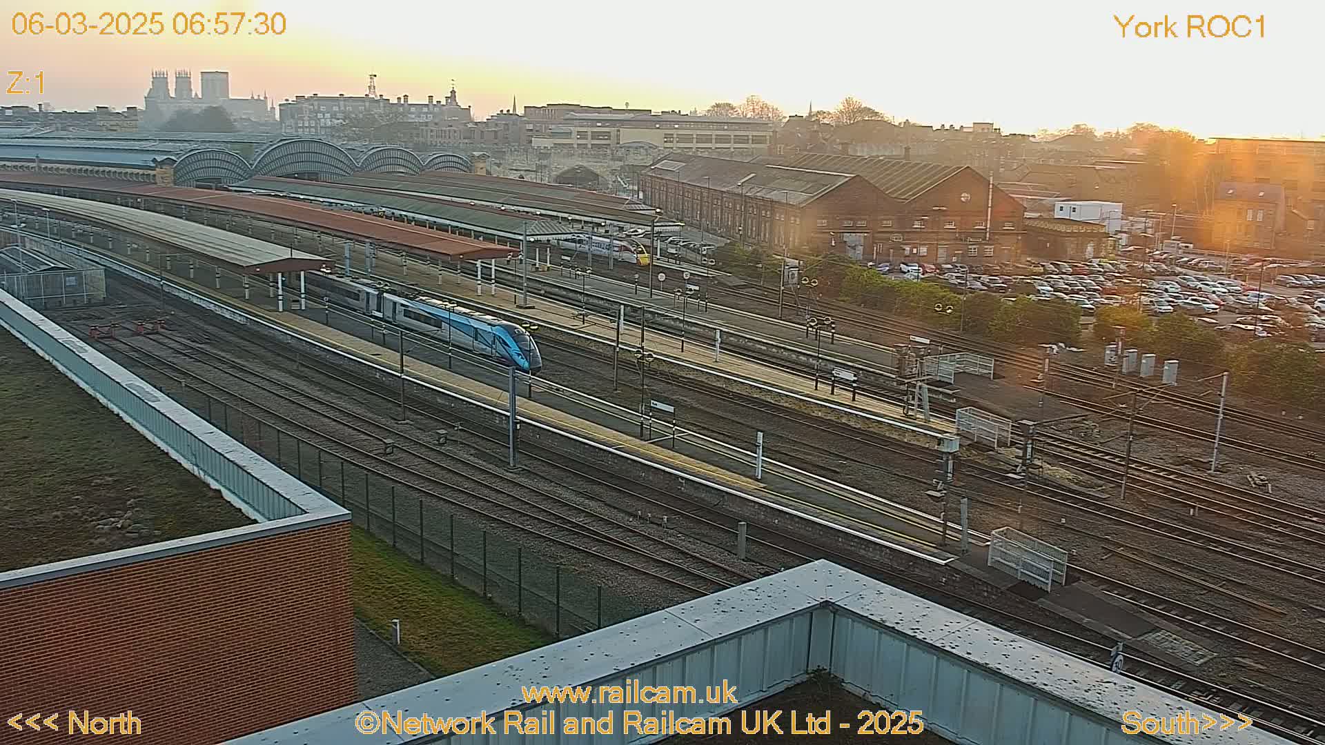 A high-angle, long shot of a train station at sunrise, showing multiple train tracks, a modern train, and a large parking lot in the background.