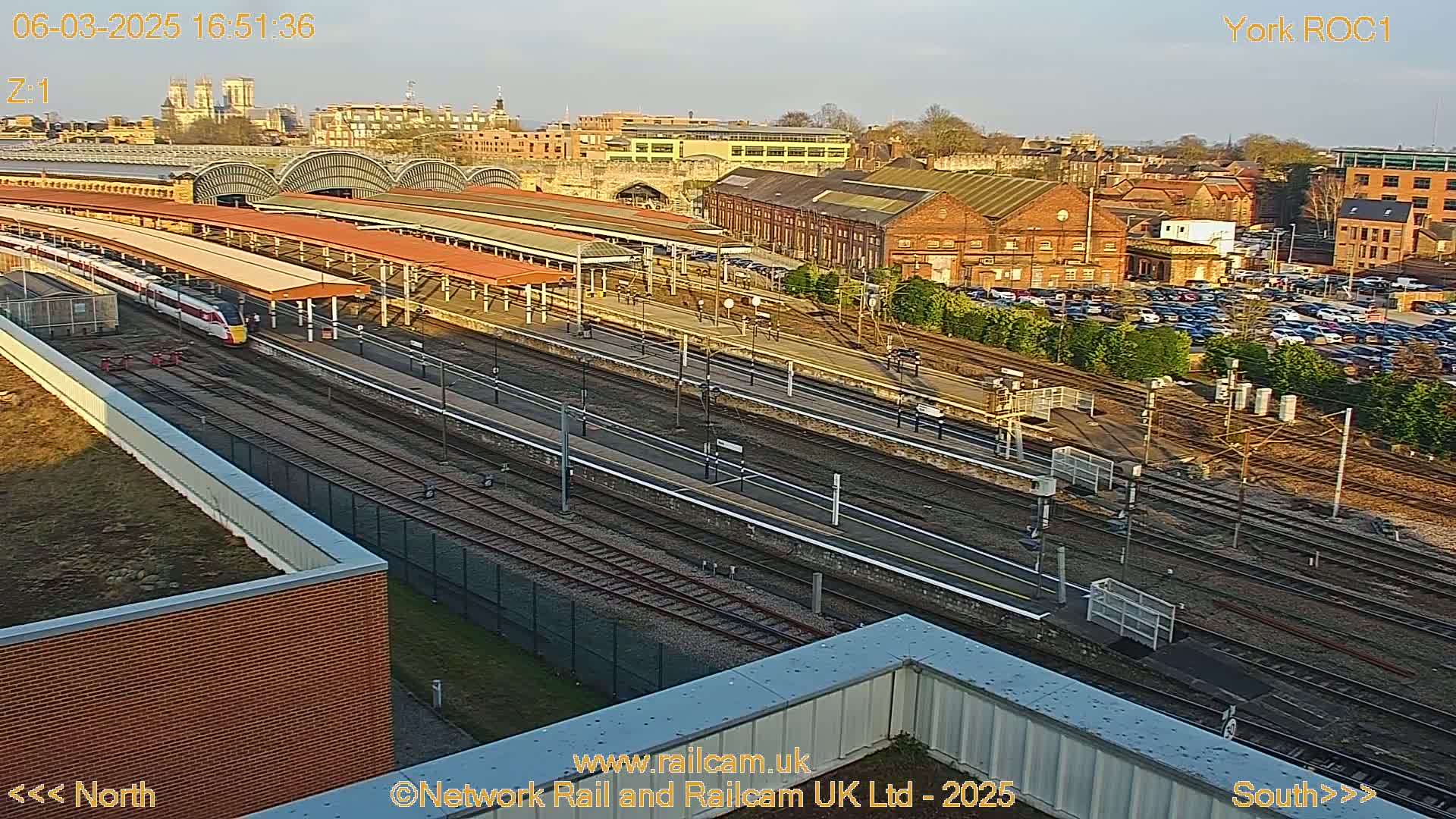 A high-angle view of a train station on a sunny day, showing multiple train tracks, platforms, and a train arriving at a platform.