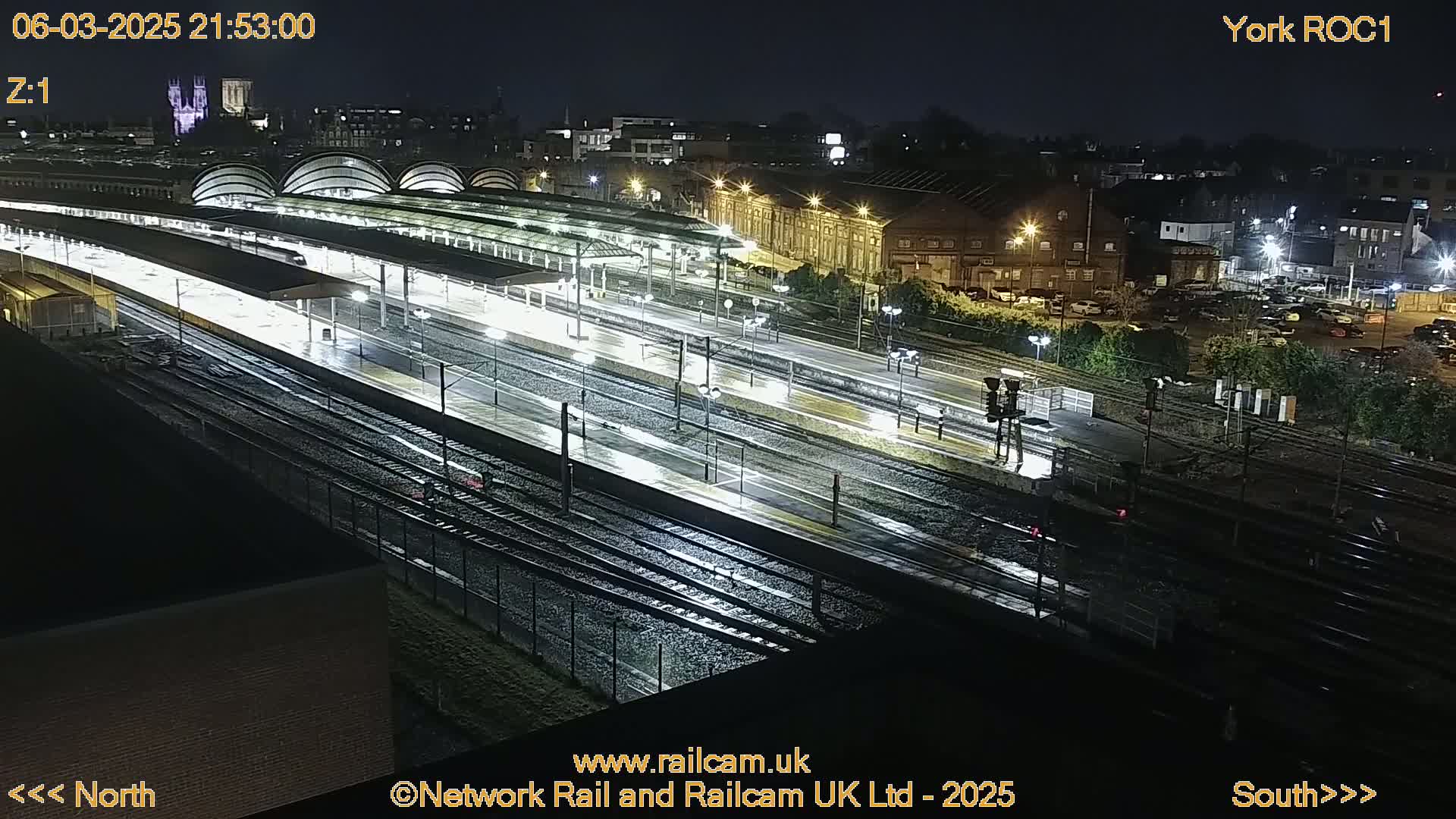 A nighttime, rain-slicked view of a train station with multiple tracks and platforms under bright lights.
