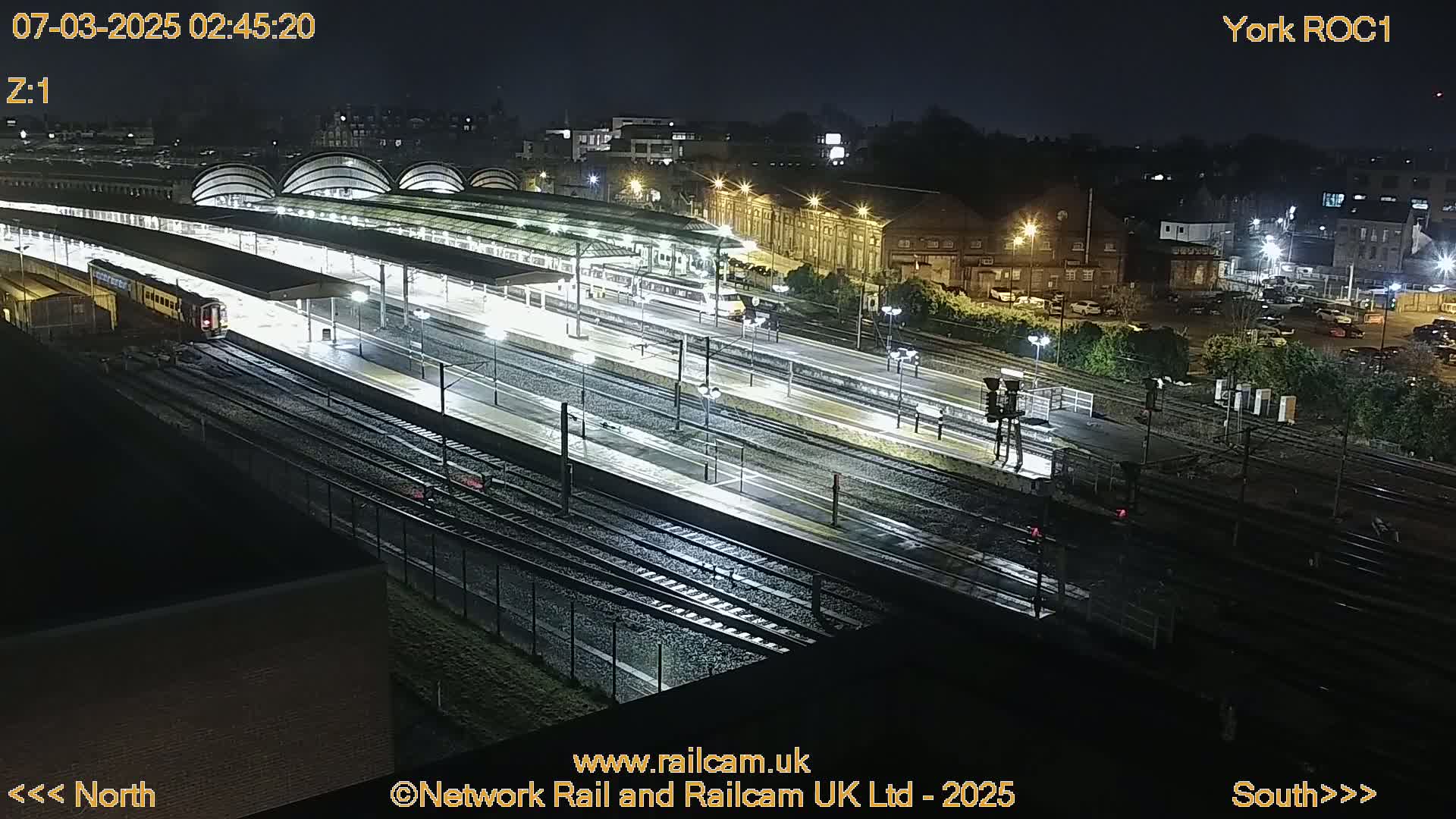 A nighttime view of a rain-slicked train station with multiple trains and tracks under illuminated canopies.