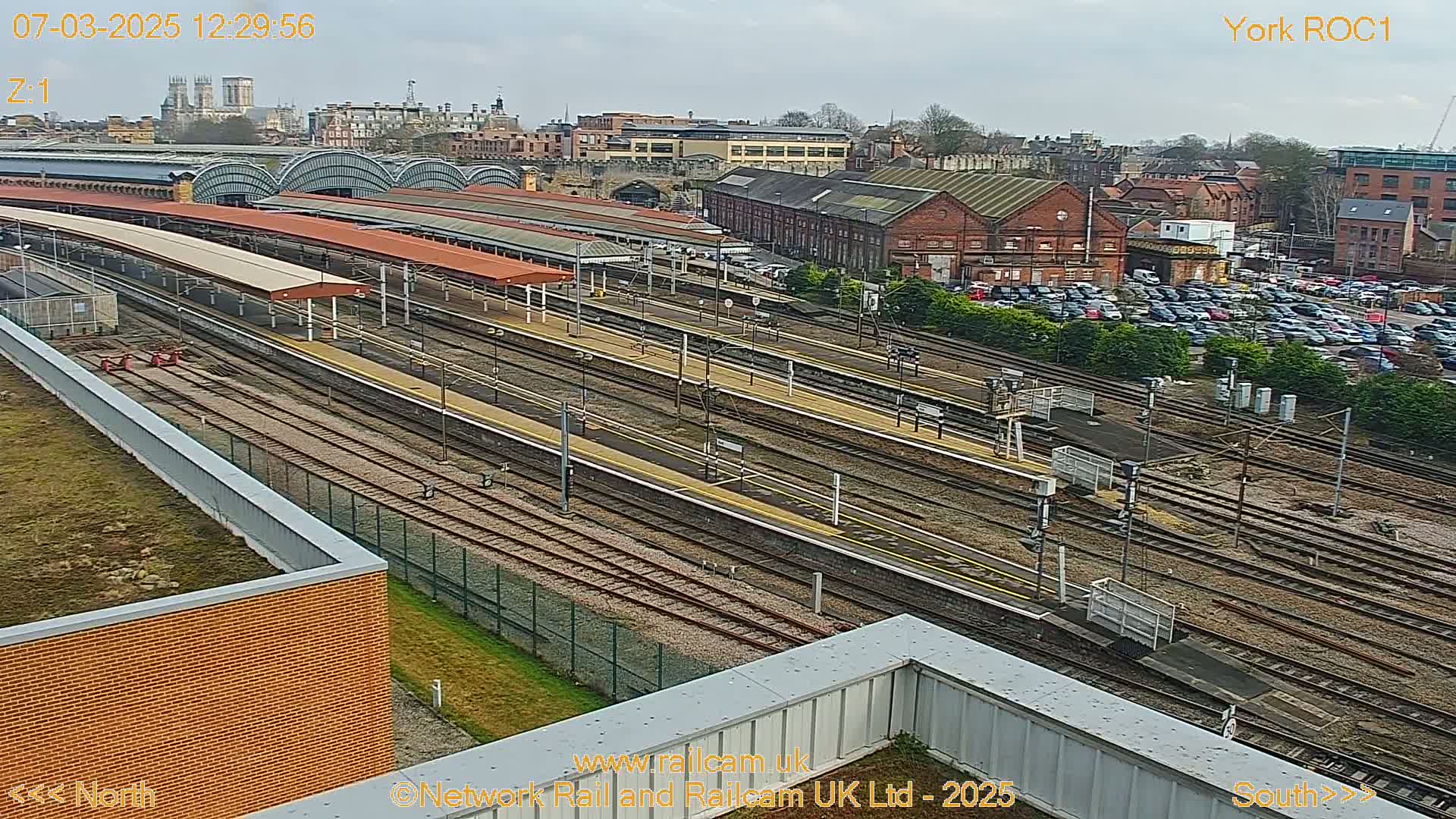 An aerial view of a train station on a partly cloudy day, showing numerous train tracks and platforms, along with a large parking lot and surrounding buildings.