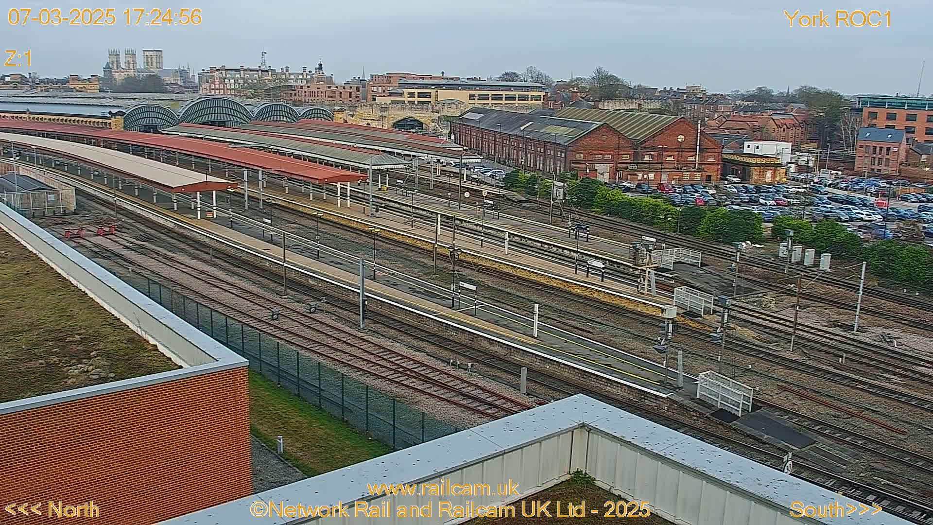 An overcast day reveals a sprawling railway station with numerous tracks, platforms, and overhead wires, surrounded by buildings and a parking lot.