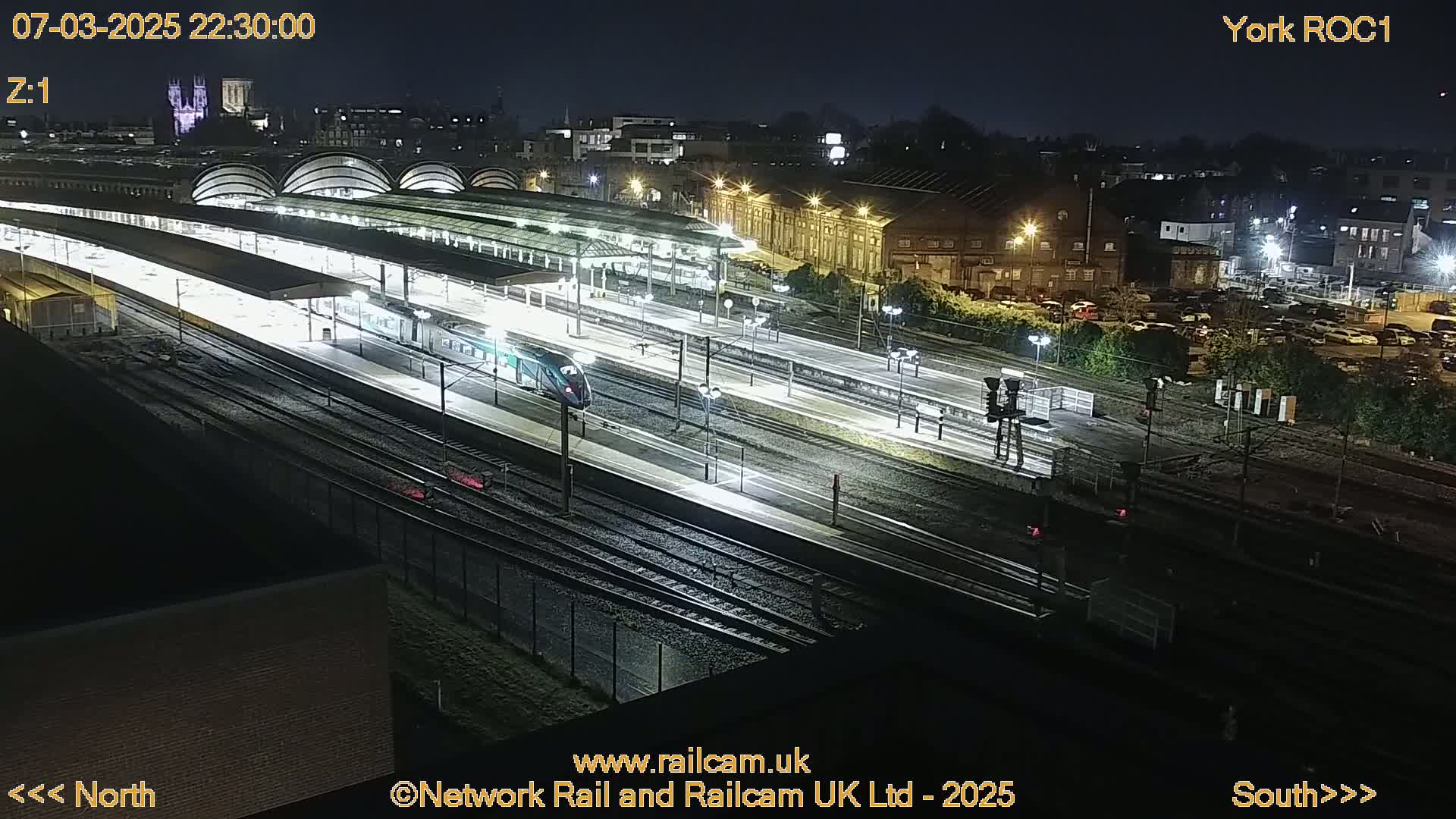 A nighttime, high-angle view of a train station with a train at a platform, surrounded by tracks and buildings under bright lights.