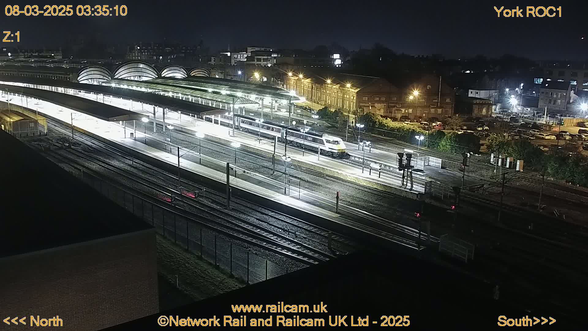 A nighttime, overhead view of a train station with a train on the tracks, surrounded by buildings and a parking lot.