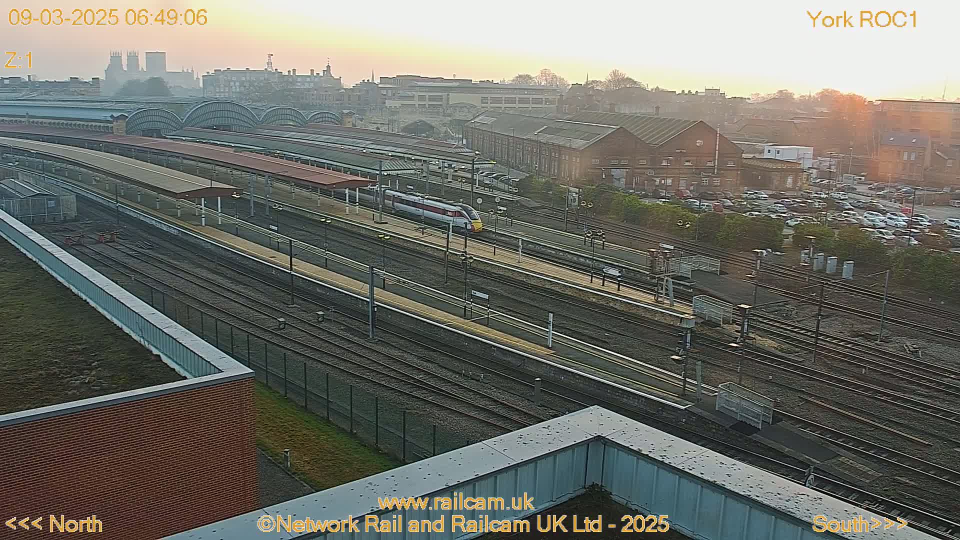 A high-angle, long shot of a train station at sunrise, showing numerous train tracks, platforms, and a high-speed train passing through.