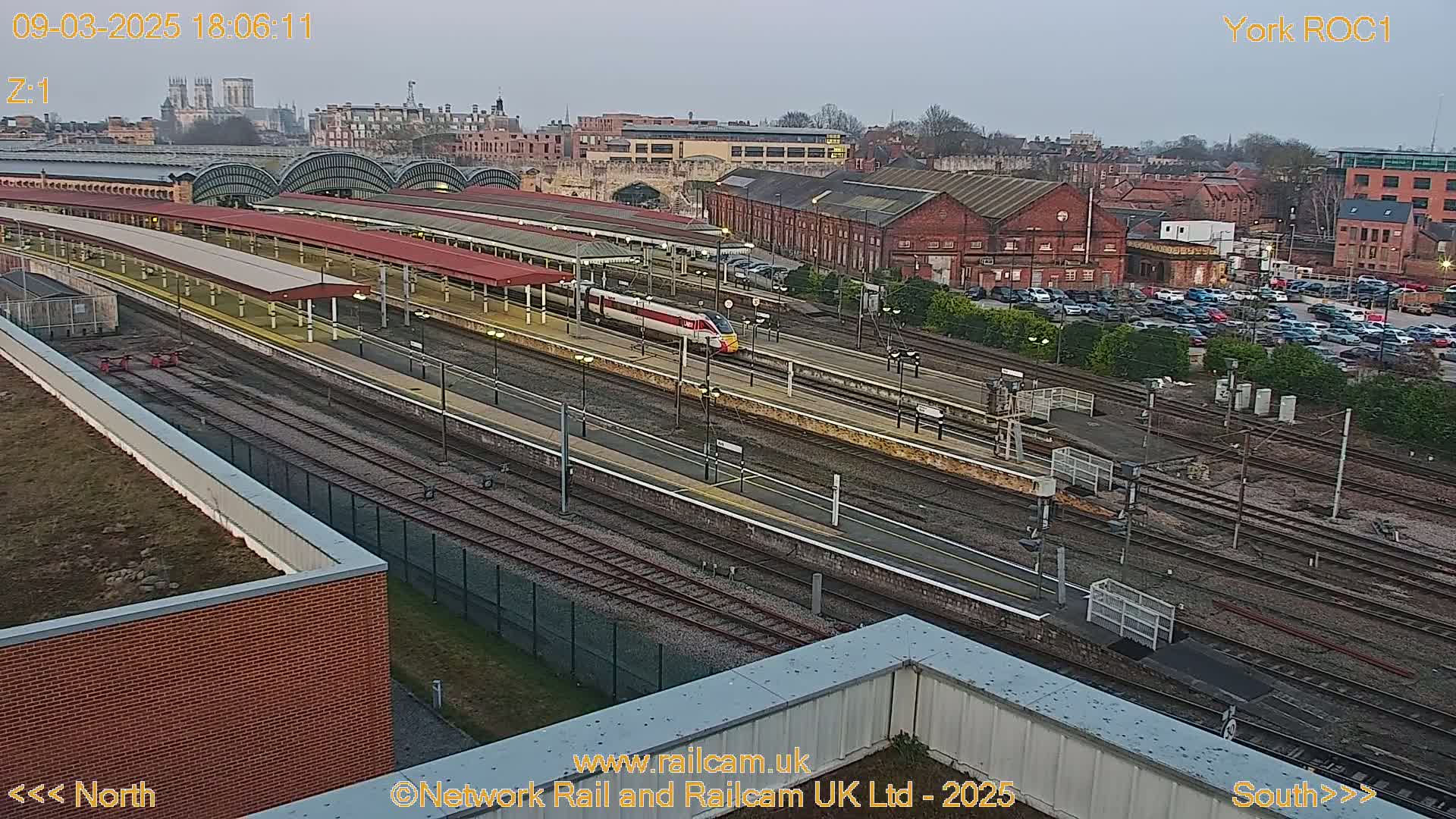 A high-angle, twilight view of a train station with a high-speed train passing through the tracks, surrounded by industrial buildings and a parking lot.