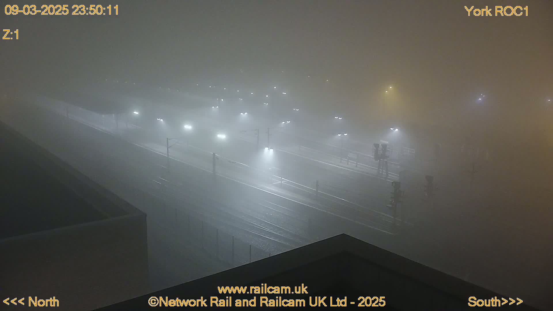 A foggy night scene shows a train yard with dimly lit tracks and vehicles barely visible in the dense fog.