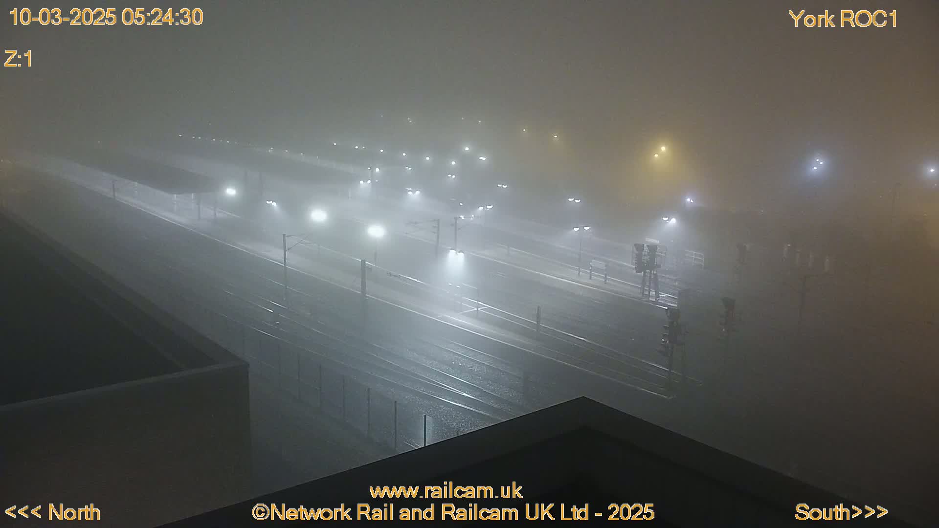 A foggy, nighttime view of a train station with numerous lights illuminating the tracks and platforms.