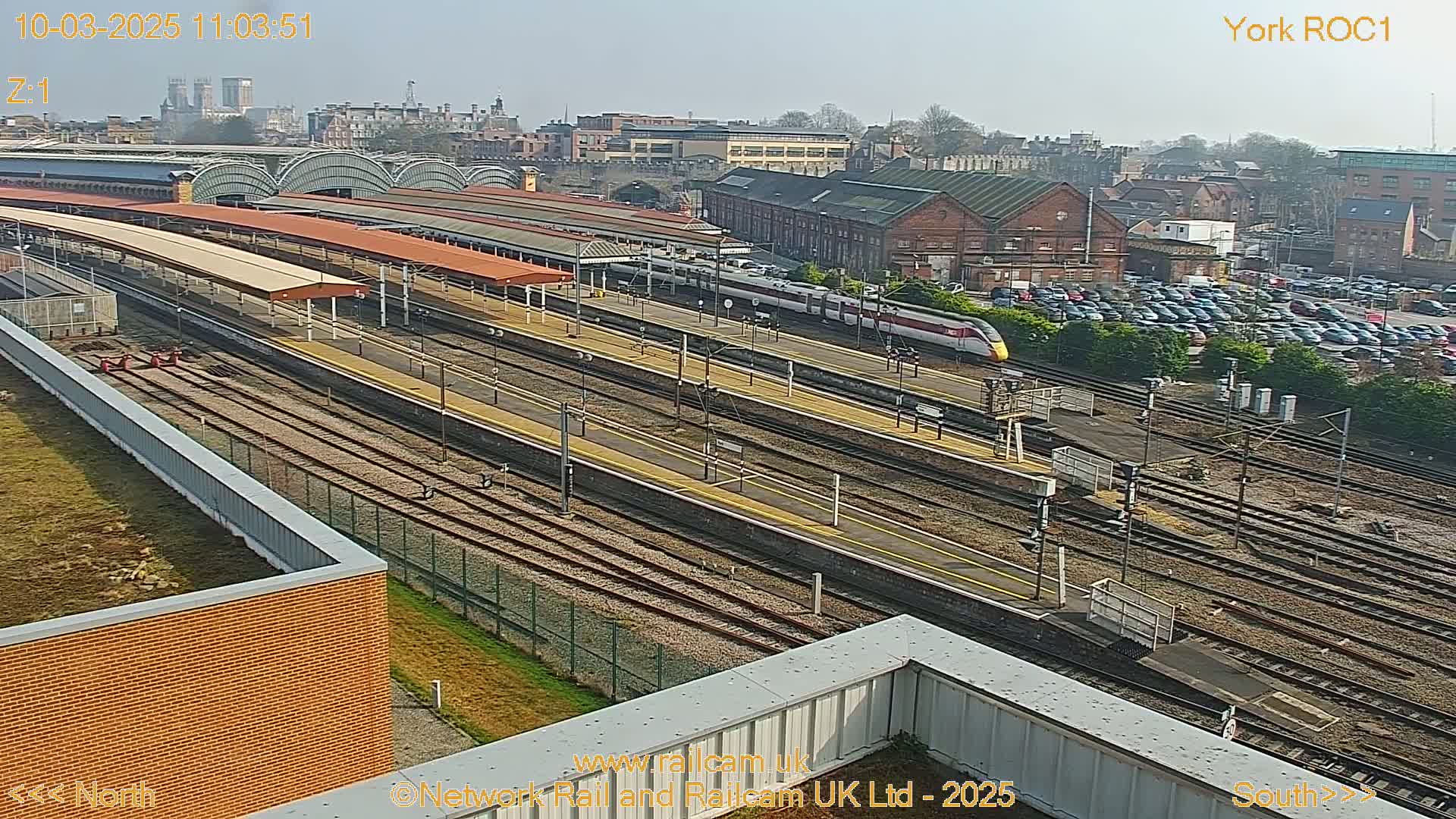 A high-angle, long shot shows a train station on a sunny day with a train passing through the tracks, surrounded by buildings and a large parking lot.