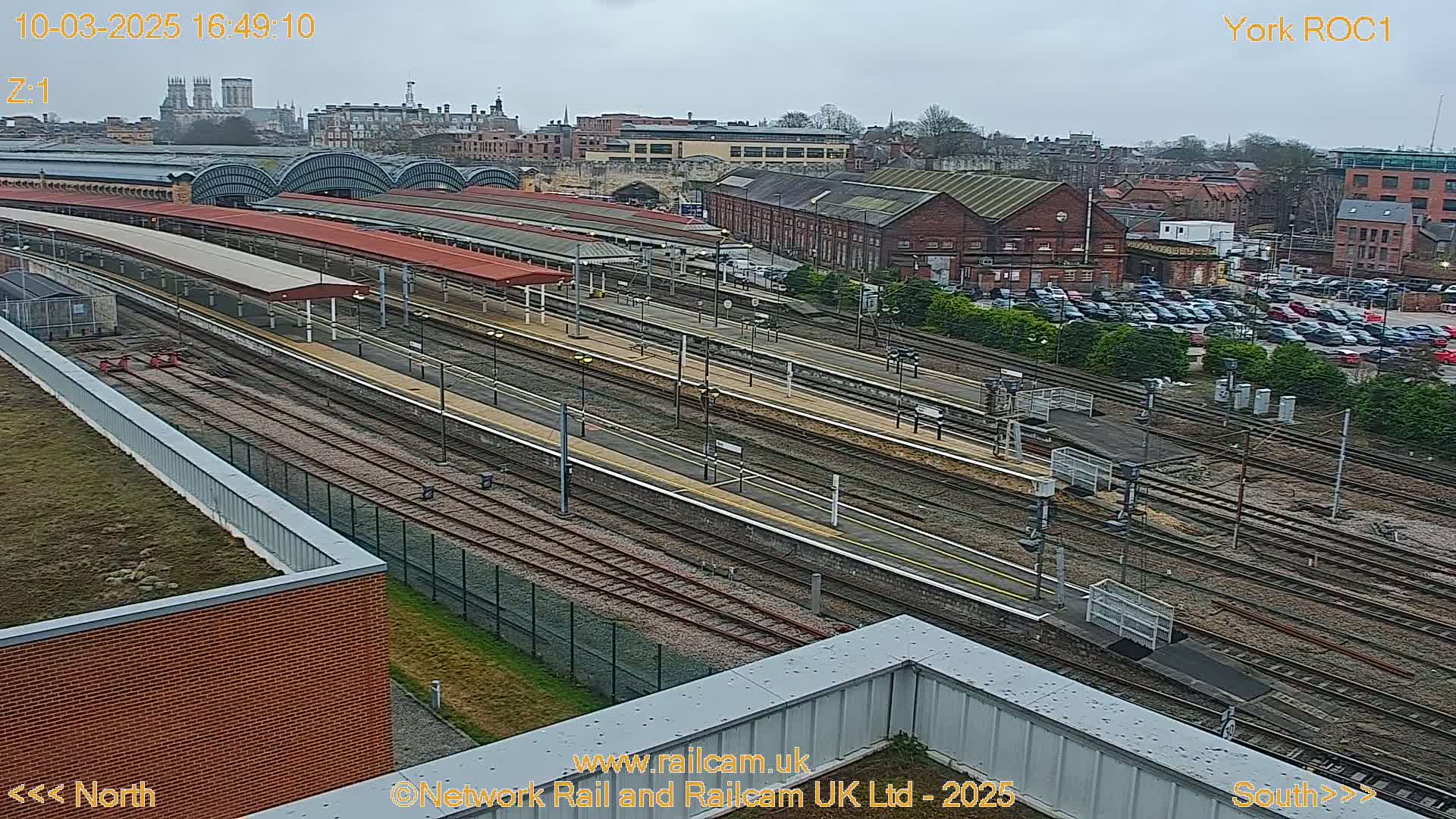 A high-angle, long shot of a train station on an overcast day, showing numerous train tracks, platforms, and buildings, with a city skyline in the background.