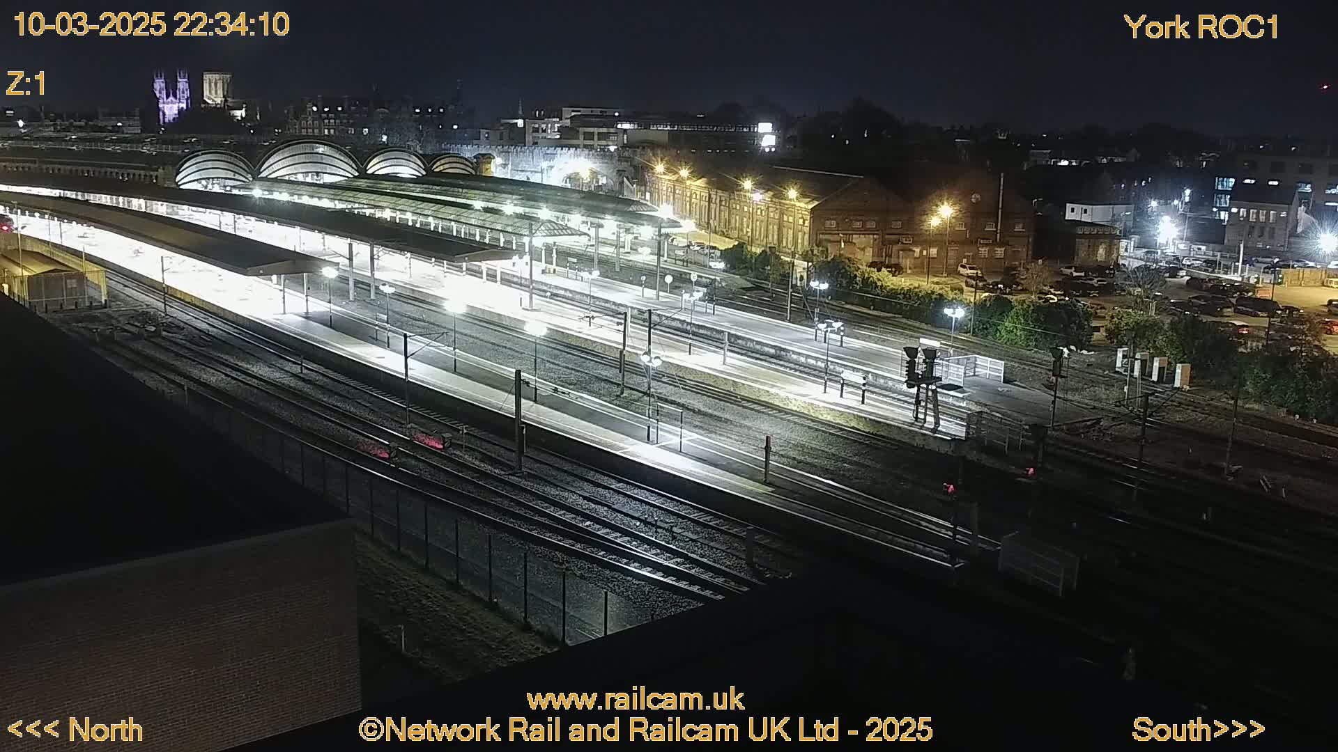A nighttime, overhead view of a brightly lit train station with multiple tracks and platforms, surrounded by buildings and a parking lot.
