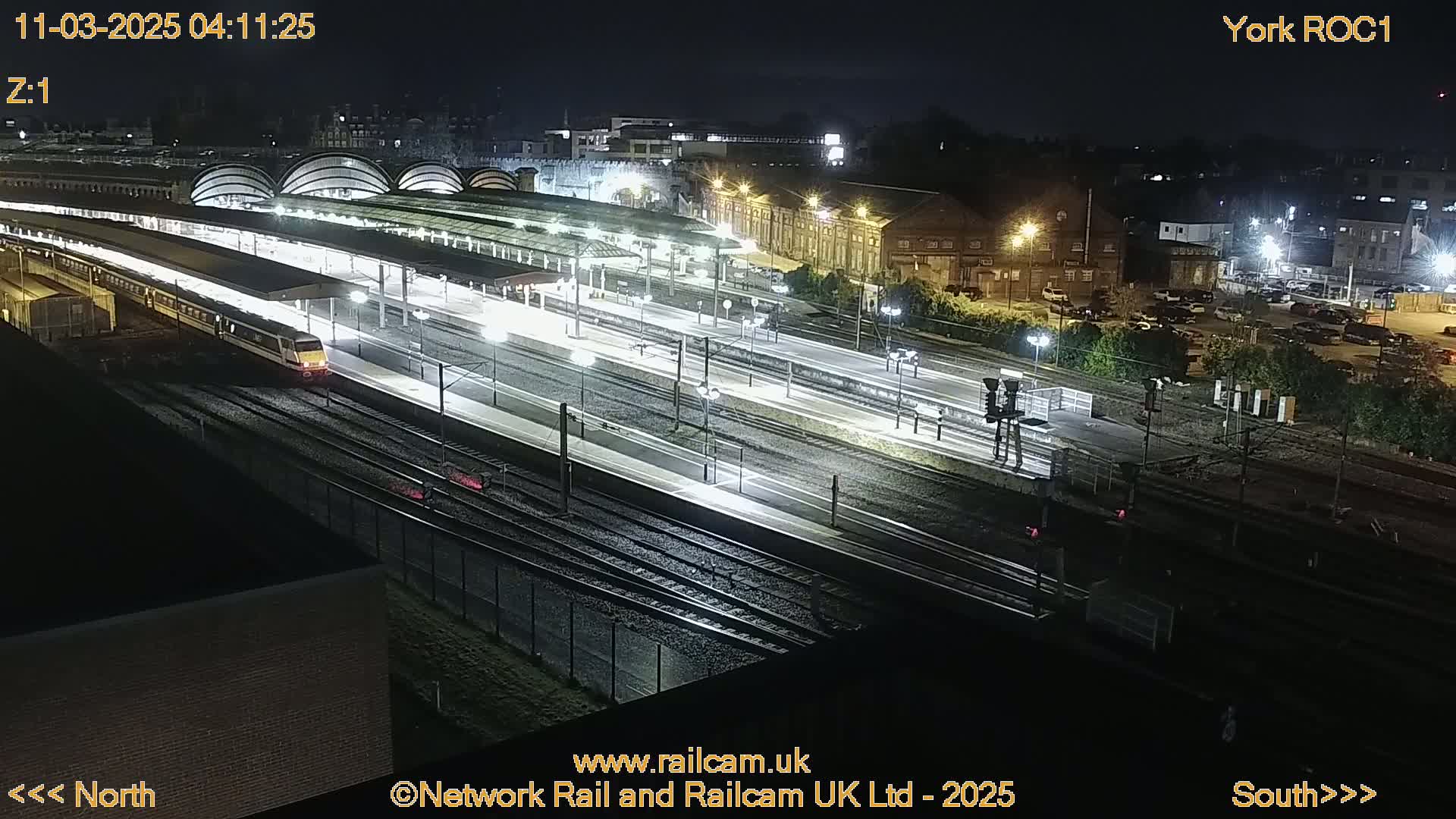 A brightly lit train station at night with a train on the tracks and several surrounding buildings.
