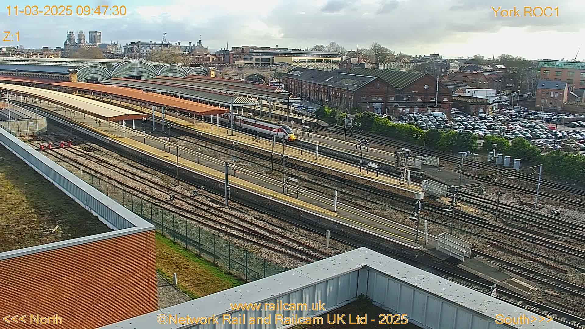 A high-angle view of a train station on a partly sunny day, showing a high-speed train passing through multiple train tracks and platforms, with a large parking lot and city buildings in the background.