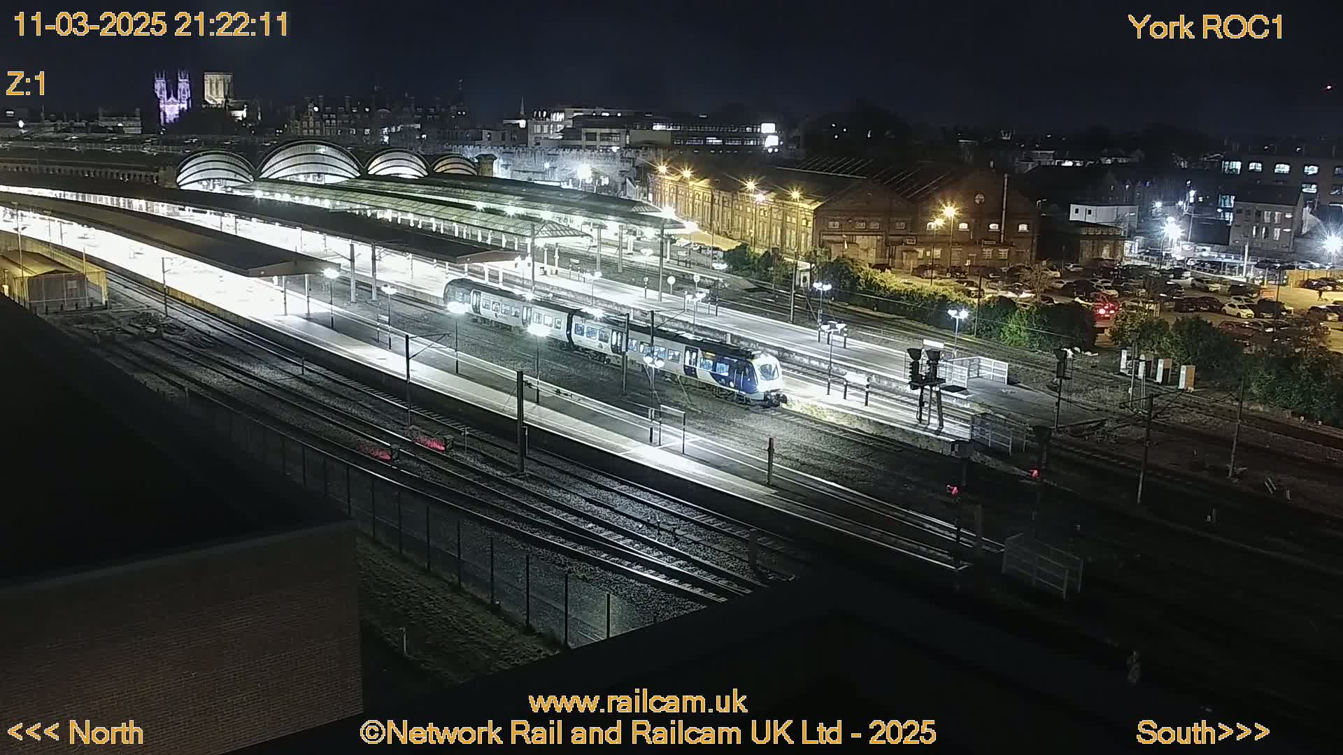 A brightly lit train station at night with a train at a platform, surrounded by tracks and buildings, under a clear sky.