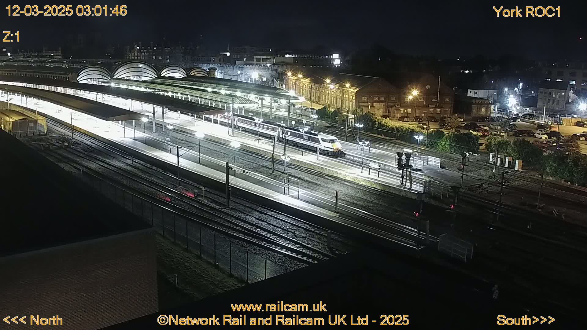 A nighttime, overhead view of a train station with a long train passing through illuminated platforms and tracks.